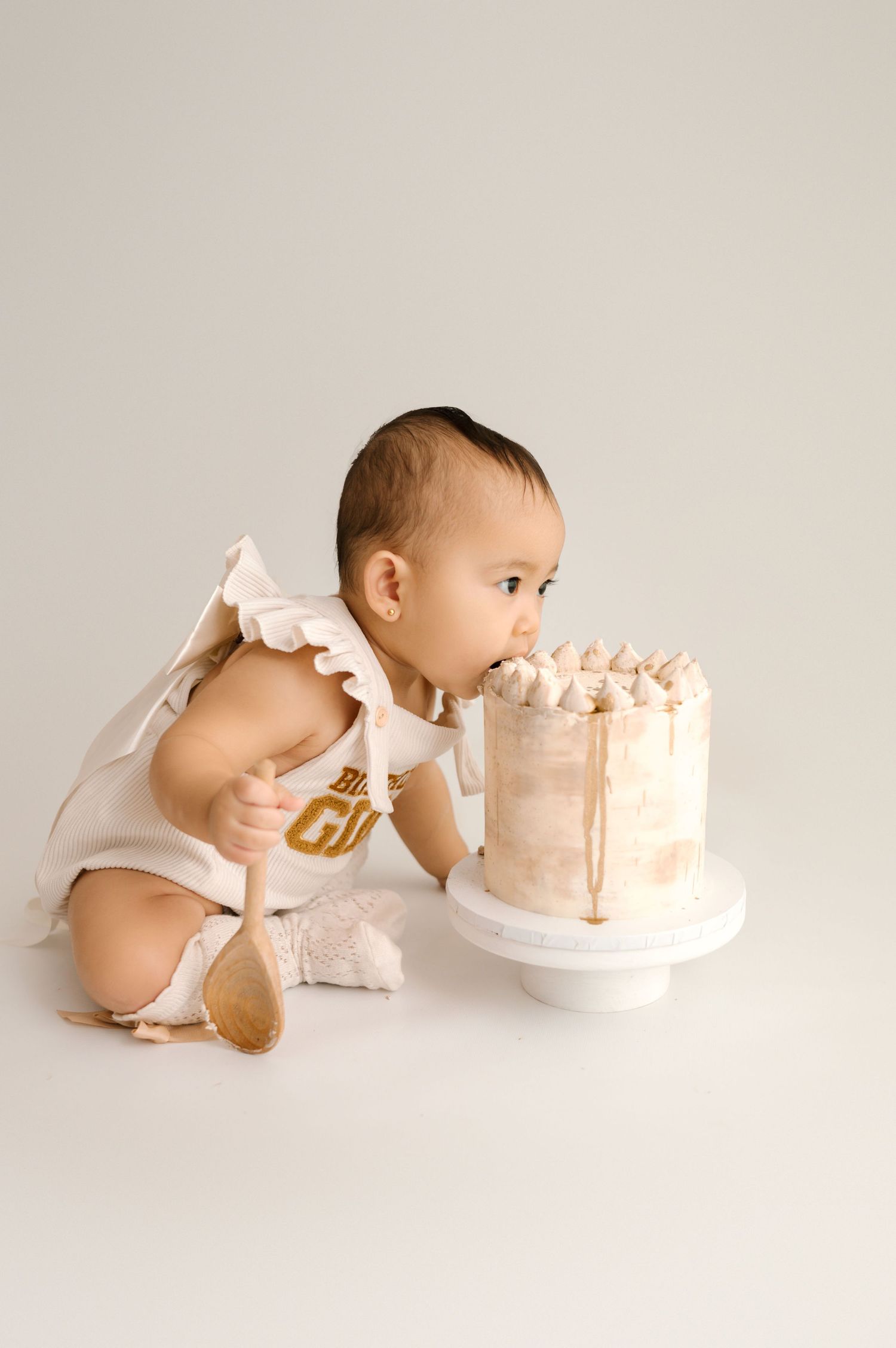 Baby in white ruffle onesie leaning into a white and gold smash cake on a cake stand.
