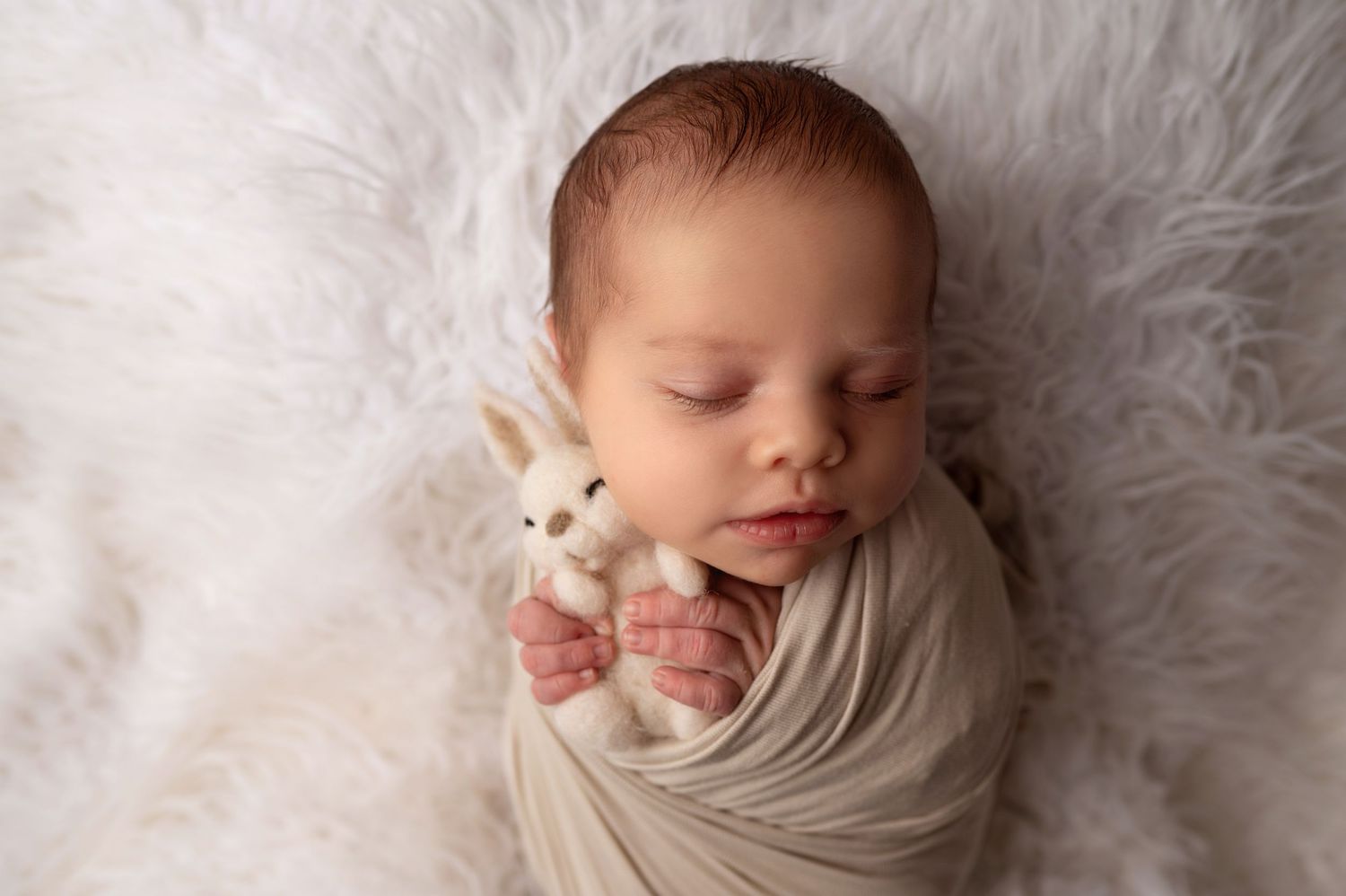 Sleeping newborn wrapped in soft white cloth with small knitted bunny toy on fluffy white fur backdrop.