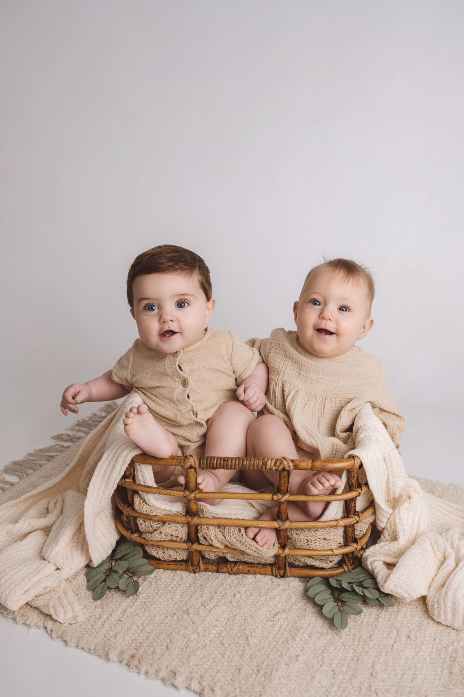 Two infants wearing beige outfits sit together in a woven basket on cream-colored fabric with green accents.
