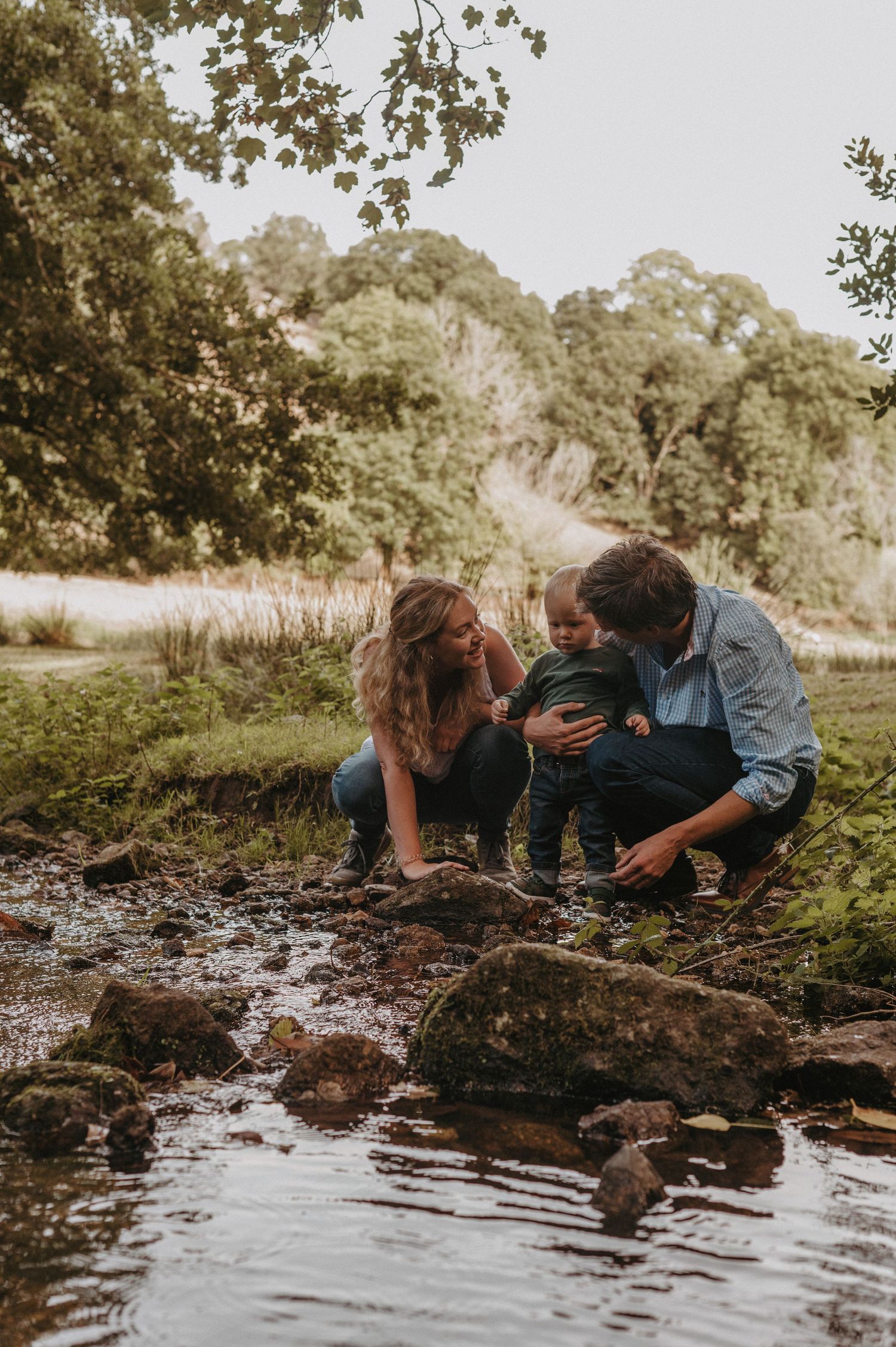 A couple and small child explore together by a stream's edge in a natural outdoor setting during golden hour.