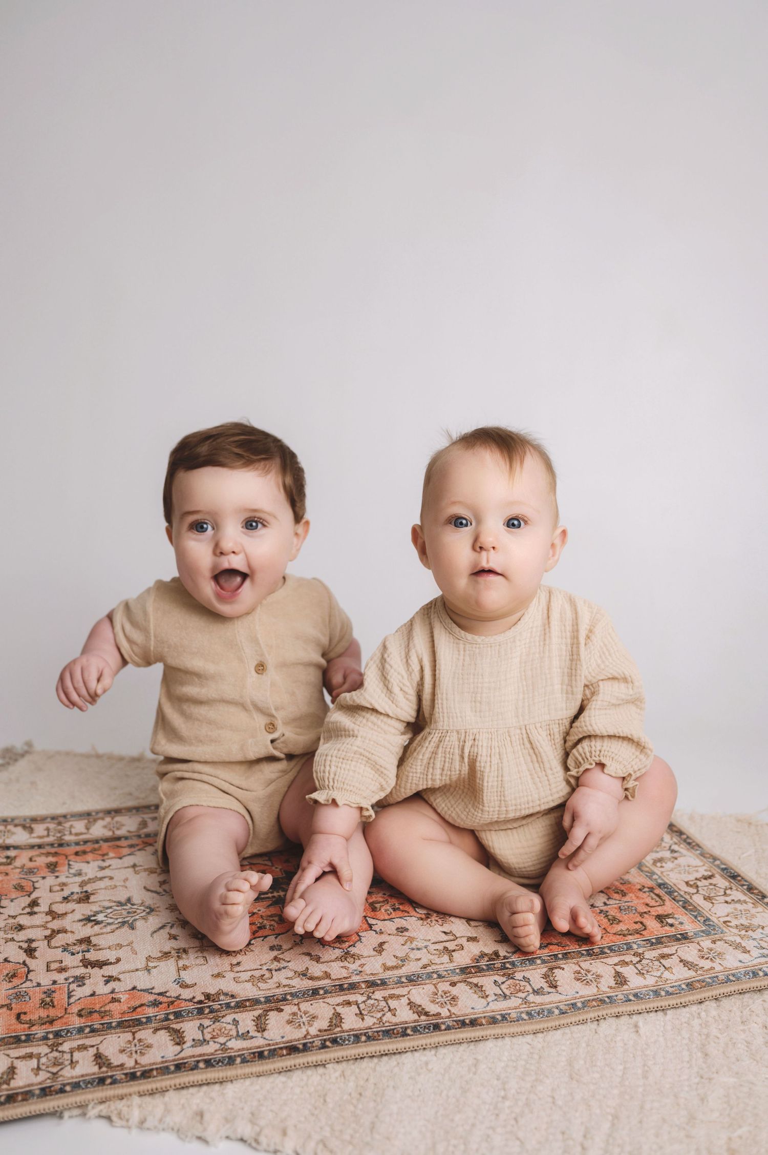 Twin babies in beige outfits sit together on a patterned rug against a light gray background.
