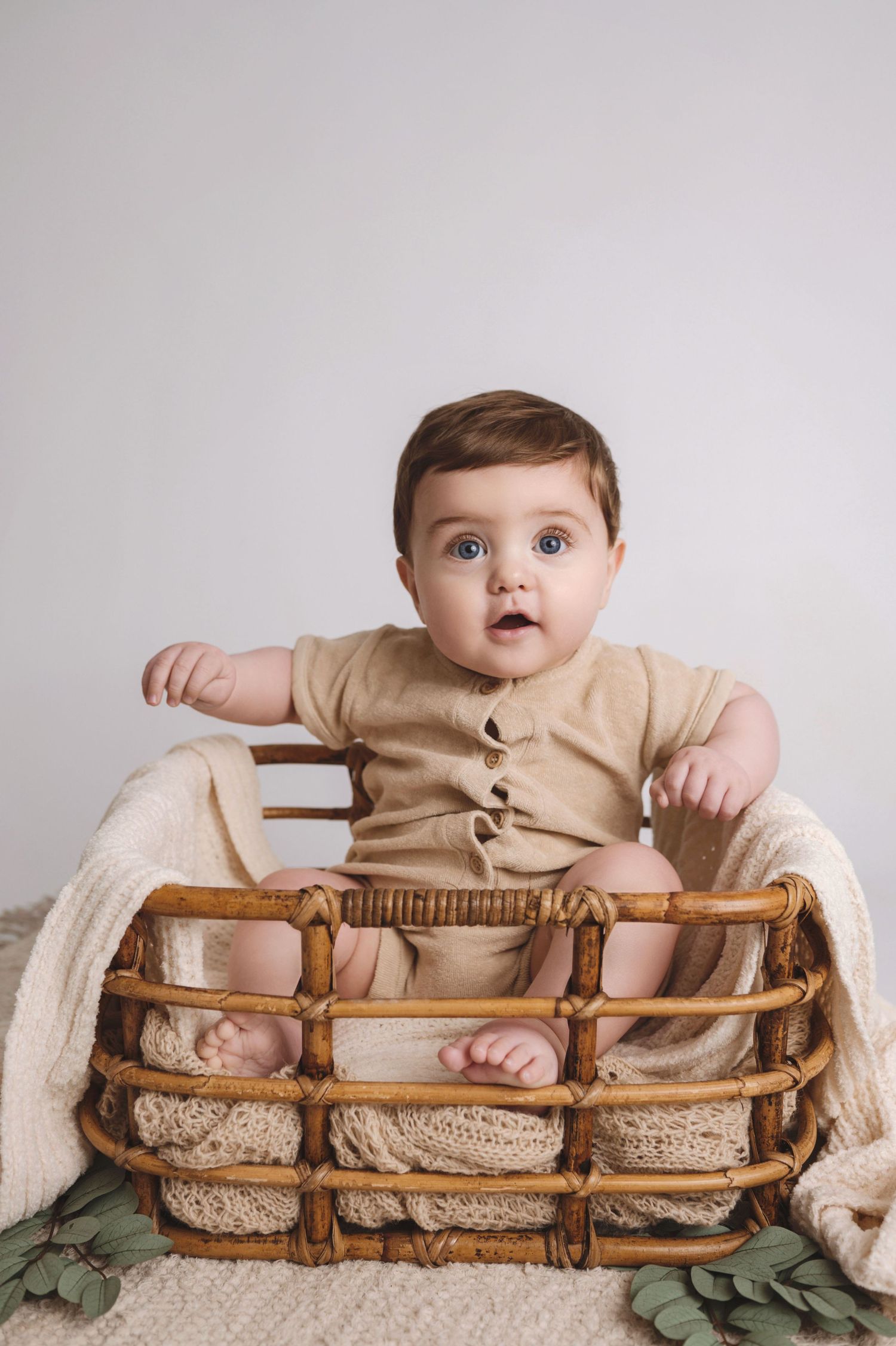 Small toddler wearing beige outfit sits in woven wooden basket with white blanket and green foliage accents.