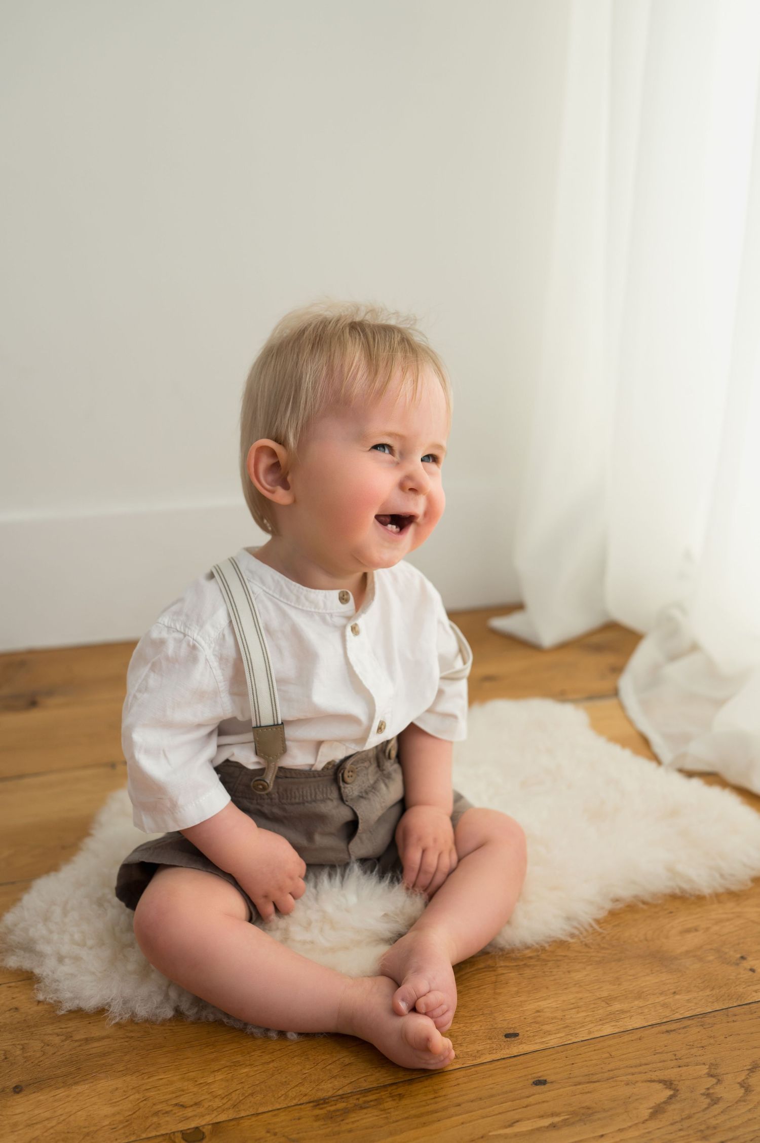 Smiling baby boy in white shirt and suspenders shorts sitting on fluffy rug on wooden floor.
