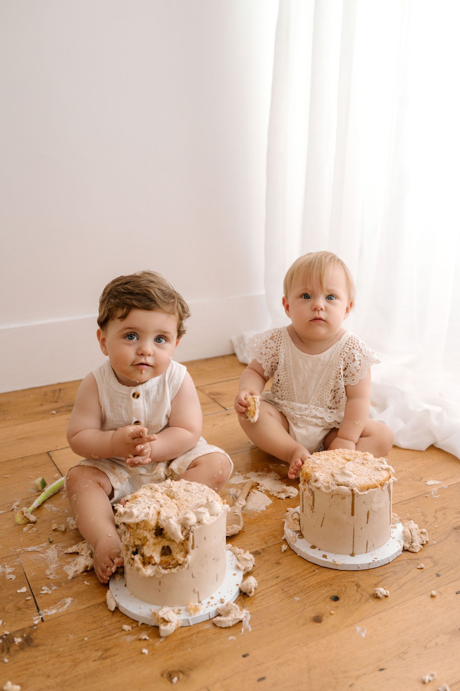 Two babies in white rompers sit on wood floor smashing their first birthday cakes in a bright studio.