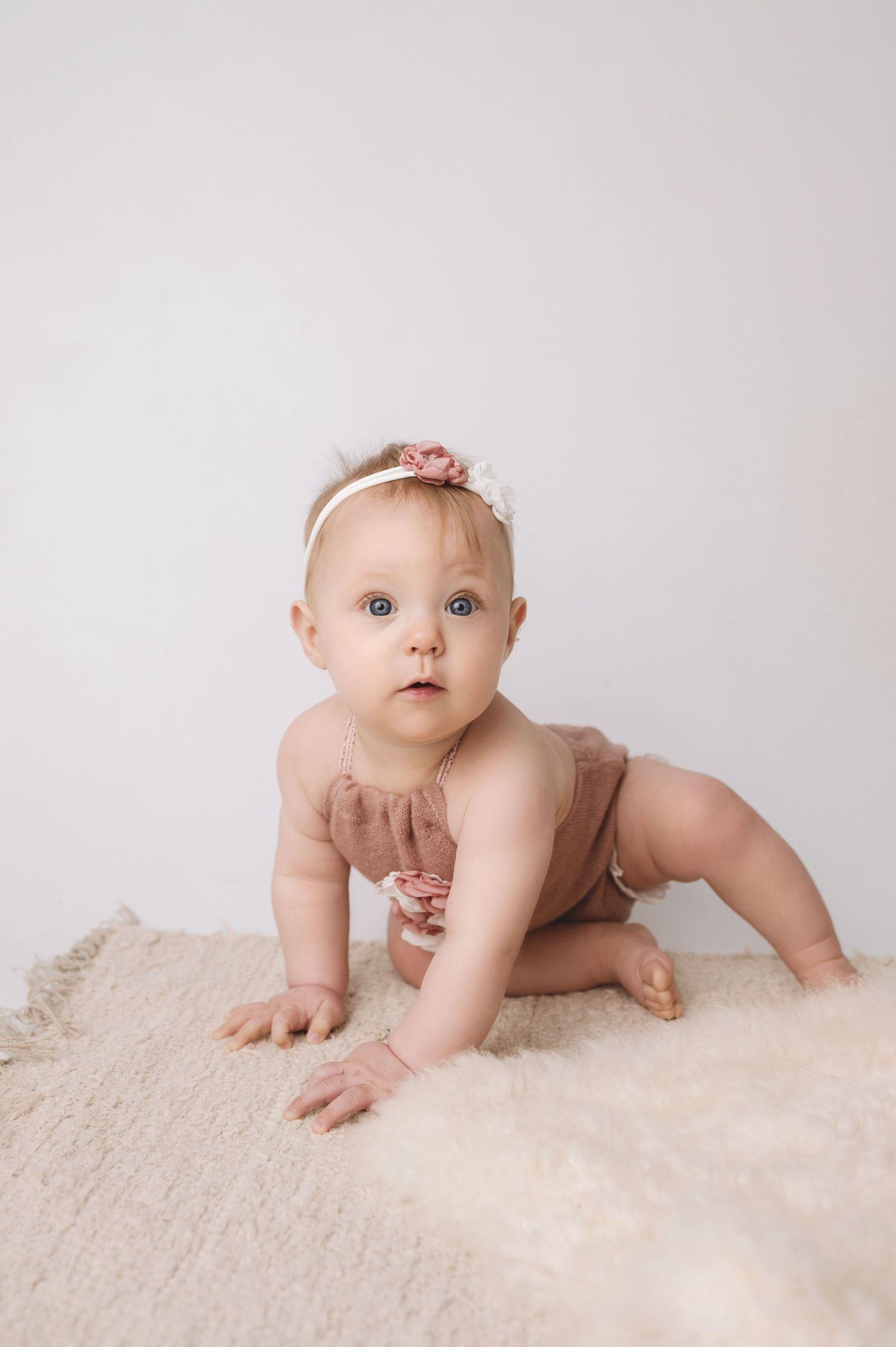 Baby wearing pink headband and ruffled outfit crawls on textured cream surface during studio portrait session.