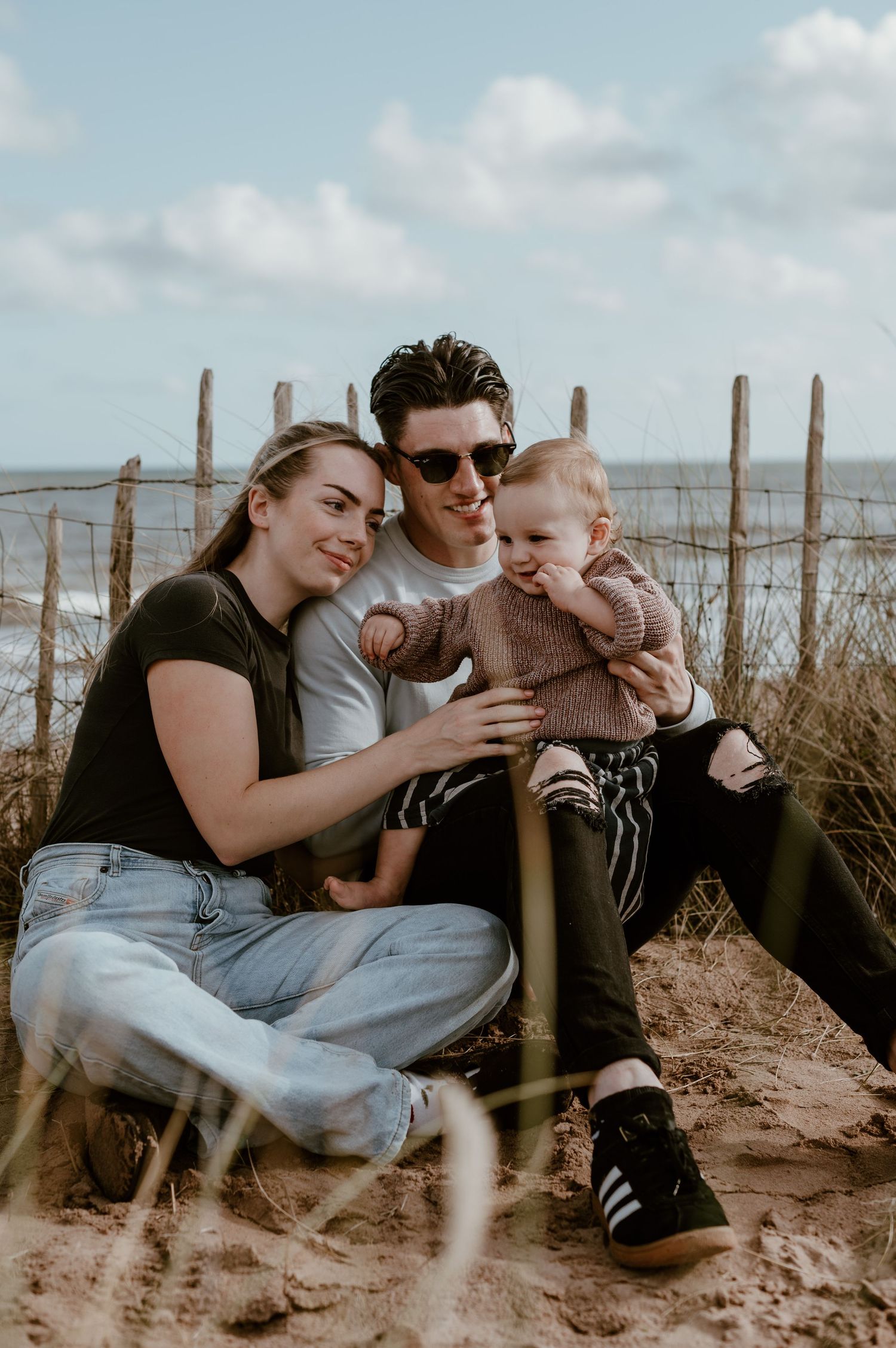 Family sitting together on sandy beach near wooden fence posts during cloudy day.