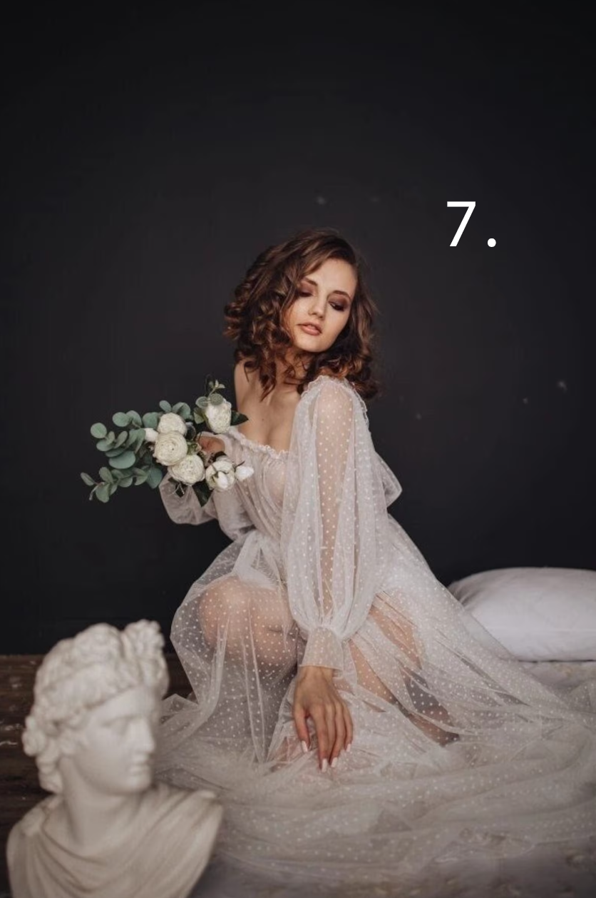 Romantic bridal boudoir photoshoot featuring delicate lace robe and white floral arrangements against dark backdrop.