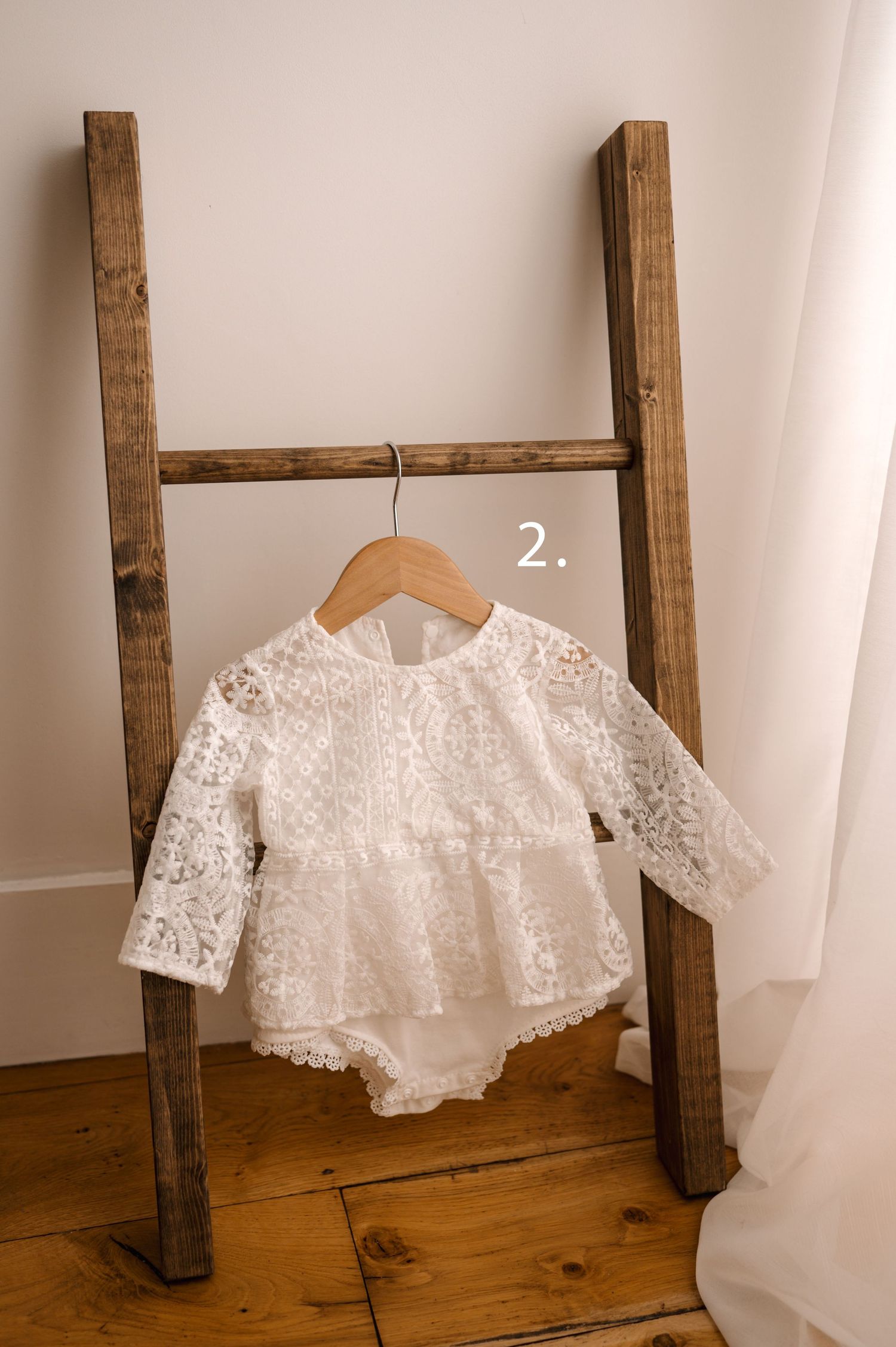 Delicate white lace bolero jacket displayed on wooden ladder against white curtain backdrop.