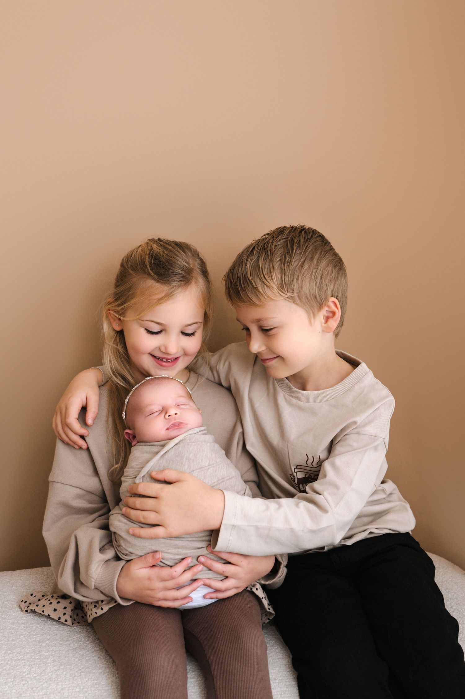 Three siblings share a tender moment together on a white surface against a neutral beige background.