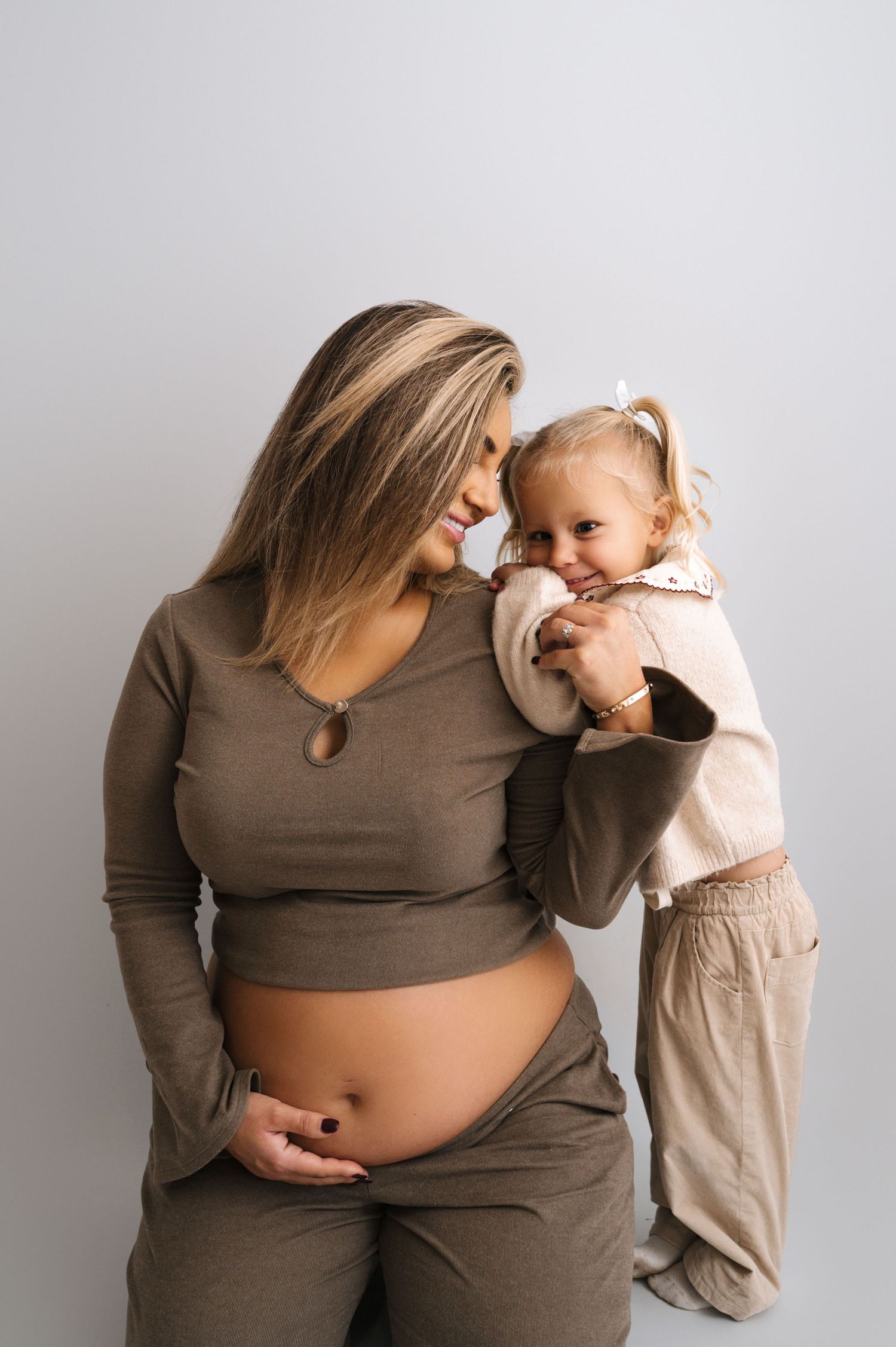 A series of maternity photos showing a pregnant woman in brown top with a small child in beige clothing against white backdrop.