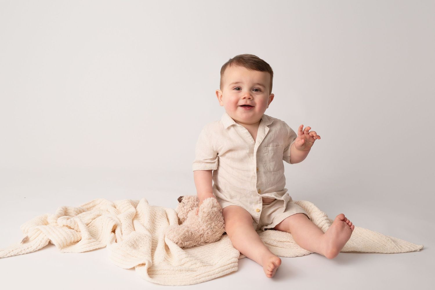 Baby in cream colored outfit sits on light beige blanket with bare feet on neutral studio backdrop.