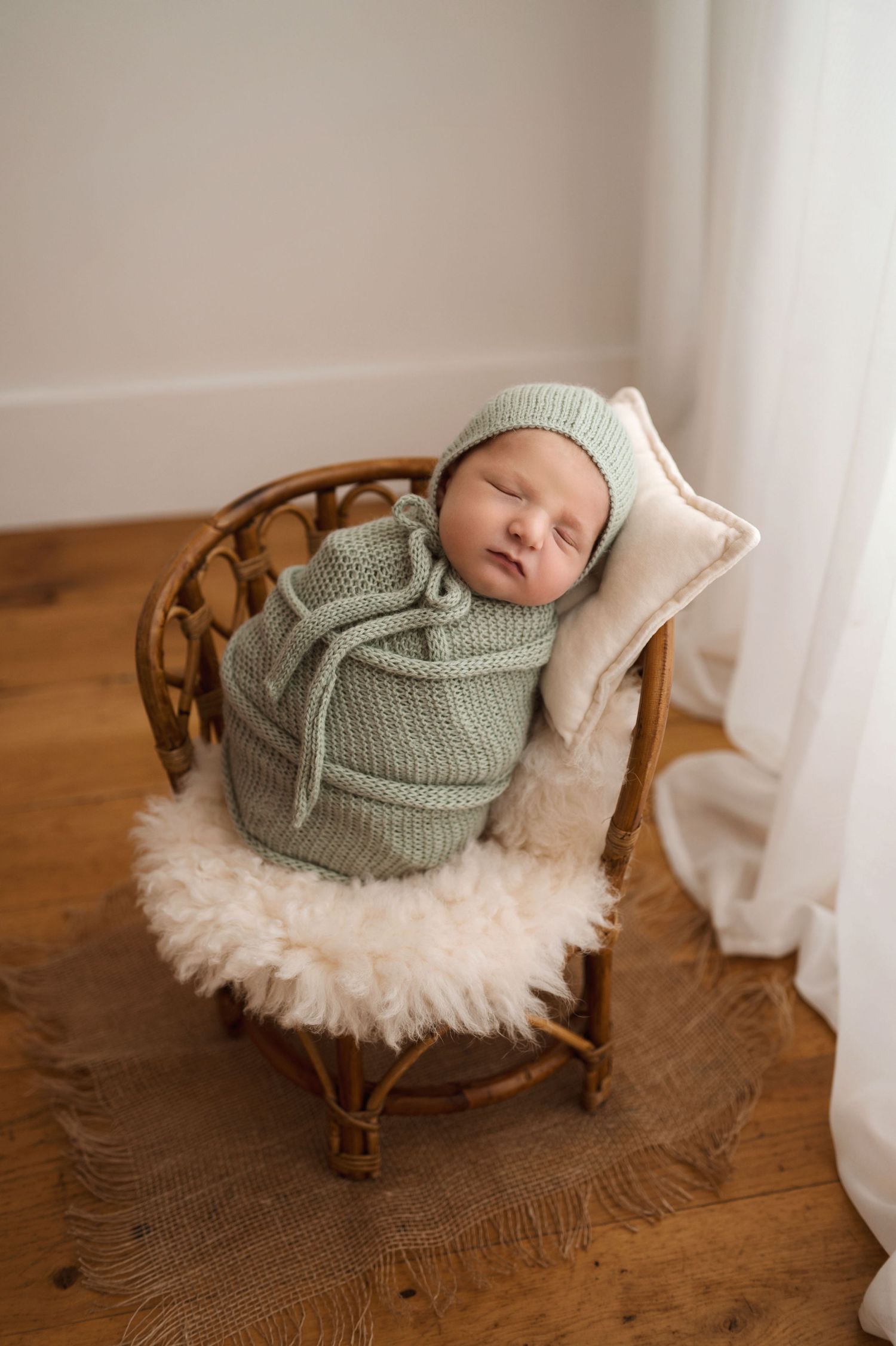 Newborn baby swaddled in sage green knit wrap and bonnet, sleeping peacefully in a tiny rattan chair with fluffy rug.