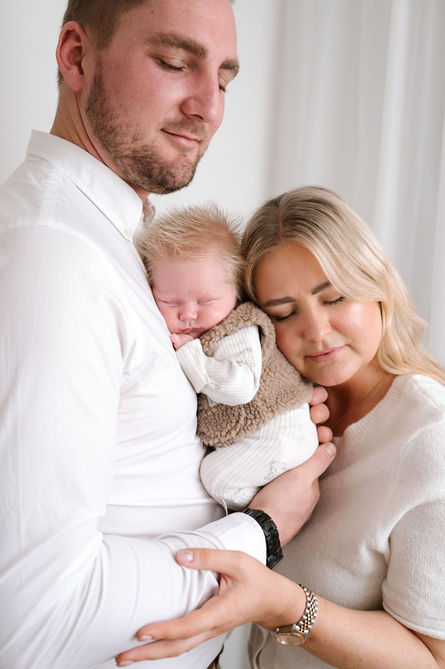 A couple tenderly holds their sleeping newborn baby, dressed in a cozy brown outfit, in a soft white studio setting.