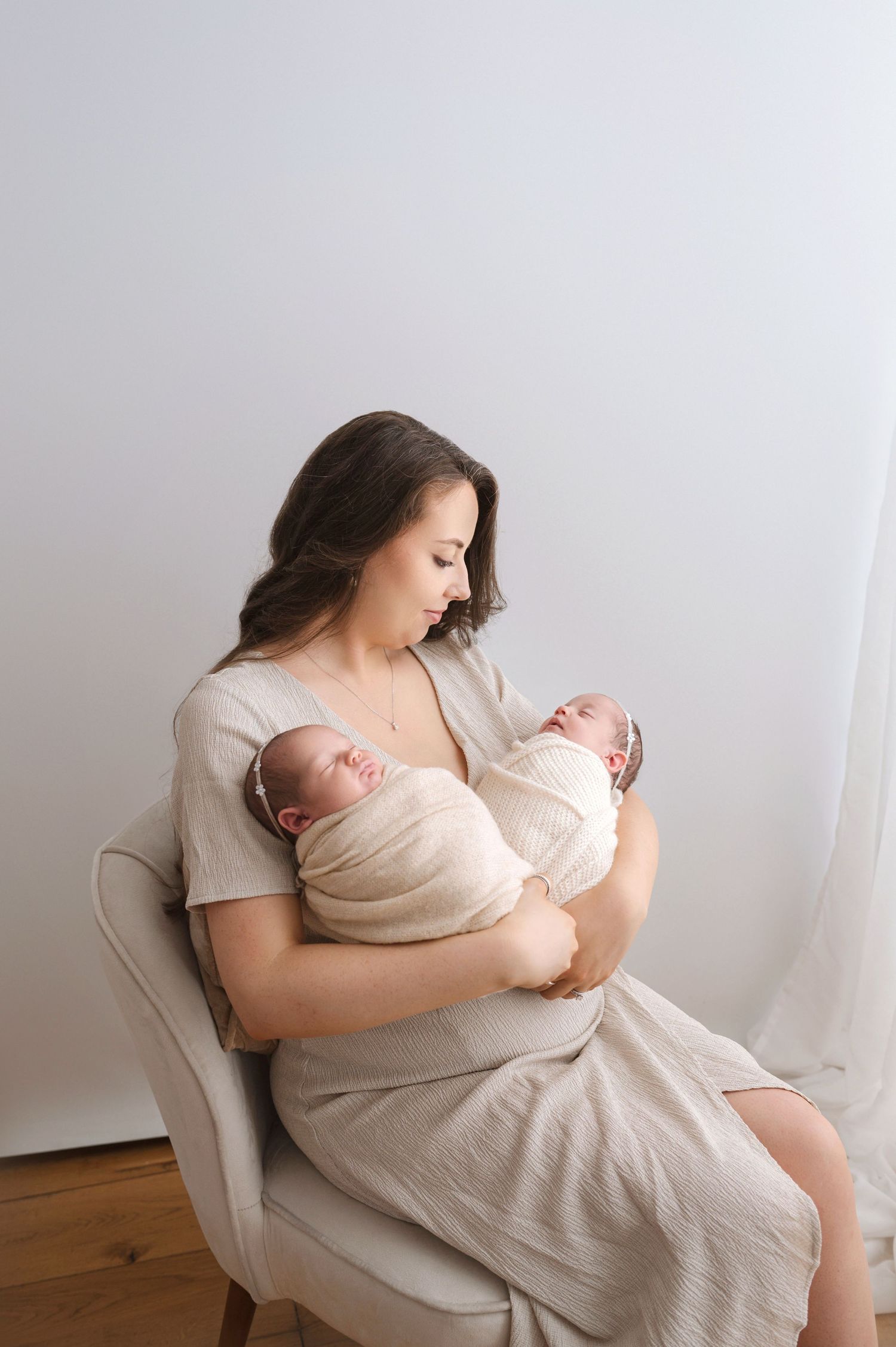 Twin newborn babies swaddled in beige wraps being cradled in an armchair during a minimalist portrait session.