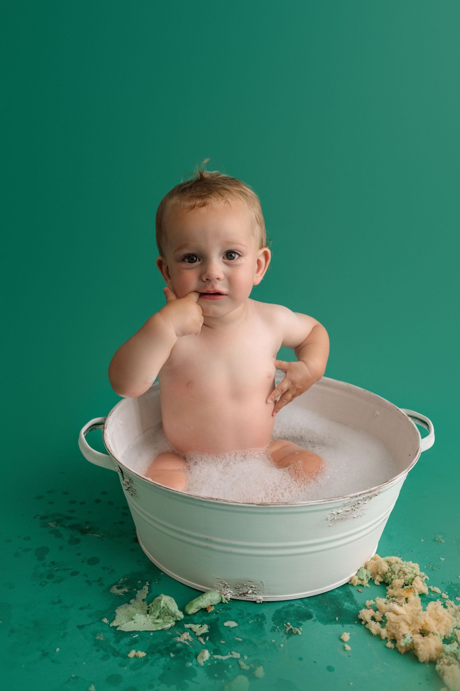 Baby sits in an antique washbasin filled with bubbles against a teal green studio backdrop.