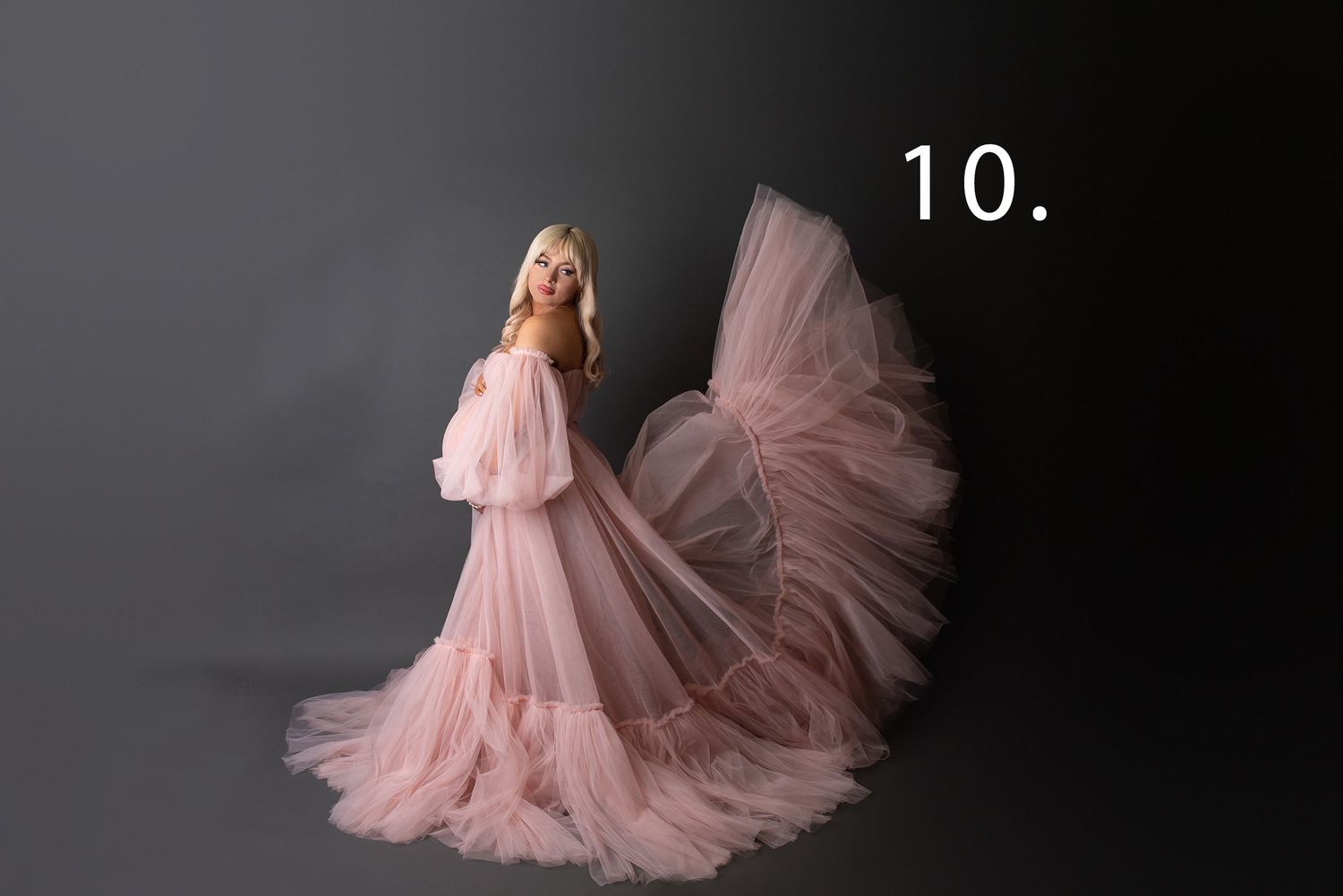 A model in a flowing blush pink tulle gown with a dramatic train poses in a dark studio setting.