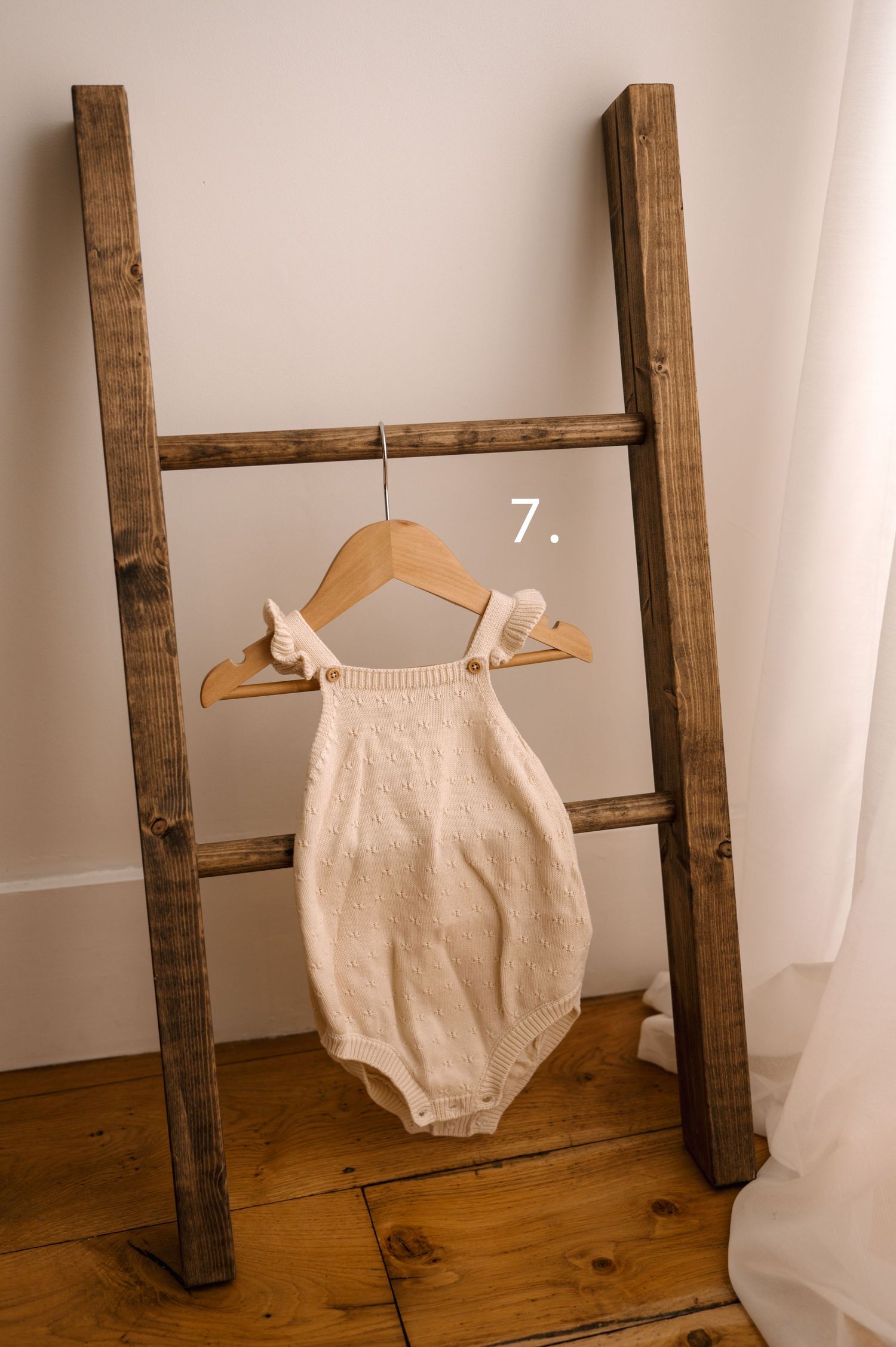 A vintage wooden ladder displays a delicate cream-colored baby romper hanging from a wooden hanger against a white wall.