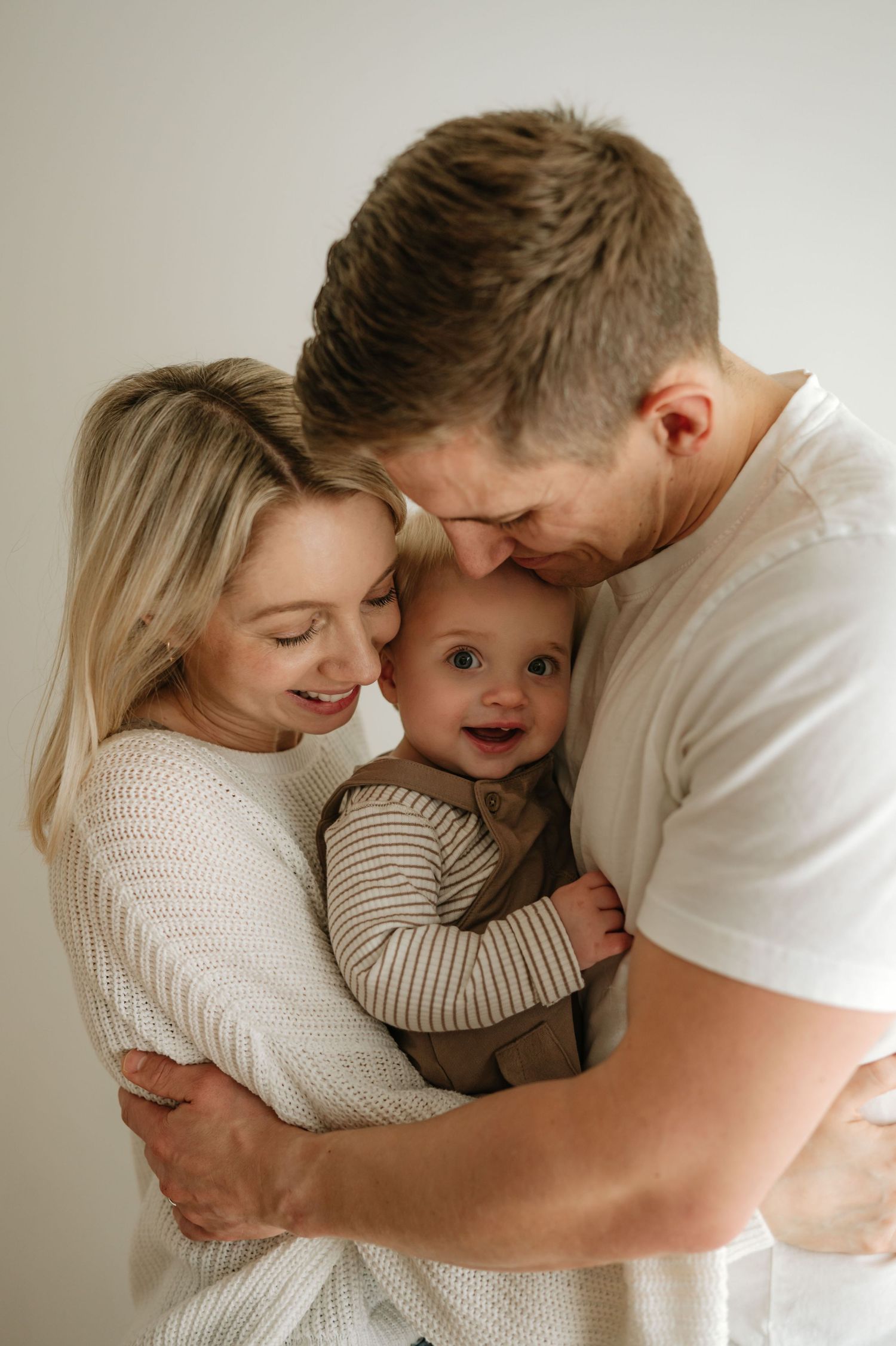 A smiling couple cuddles their happy baby boy in a warm, neutral-toned family portrait photo.