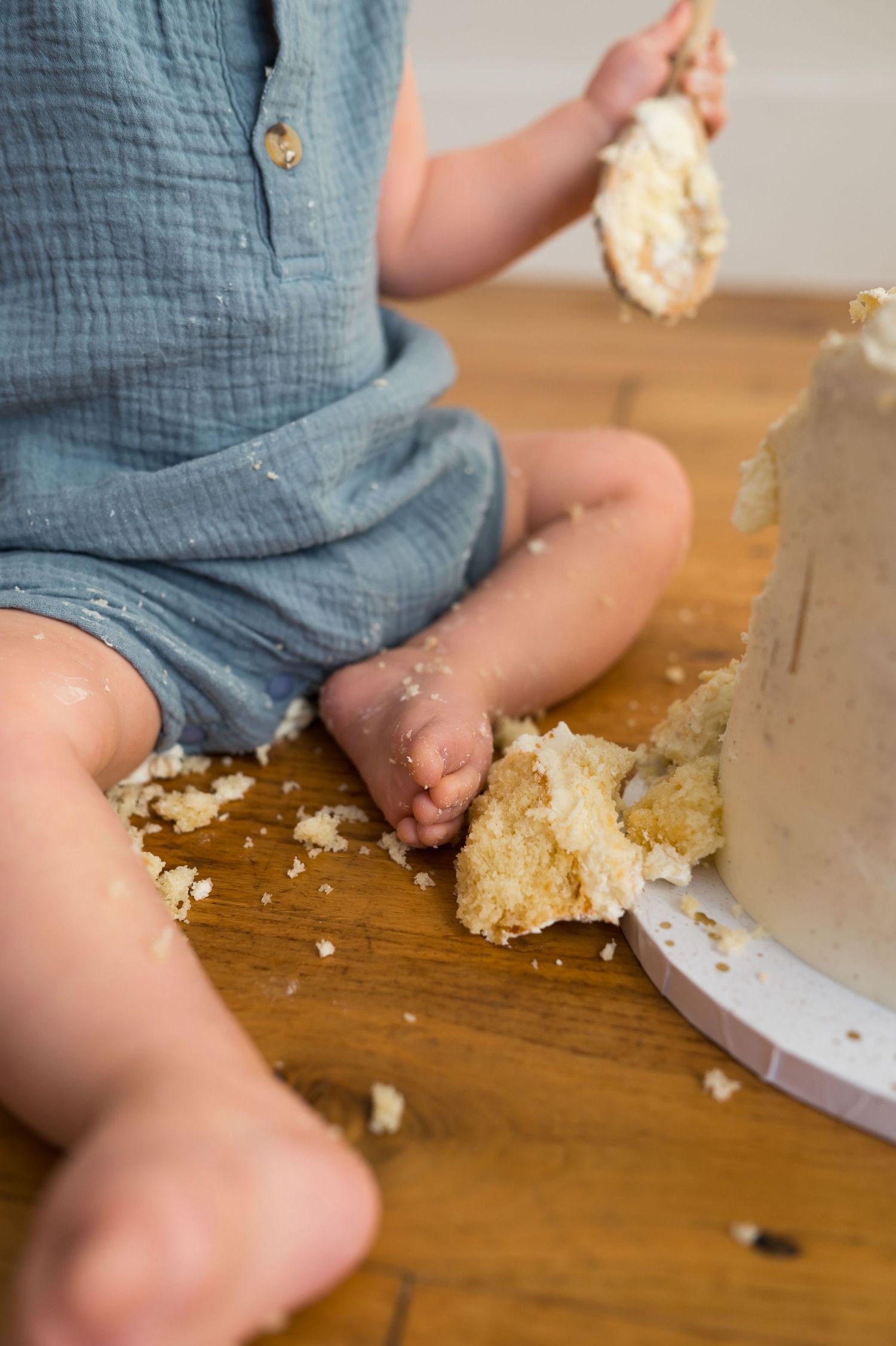 Toddler in blue romper sitting on wood floor smashing white birthday cake with hands during cake smash.