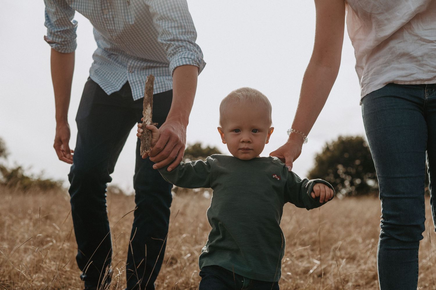 Close-up shot of a toddler in a green sweater walking while holding hands during sunset in a field.