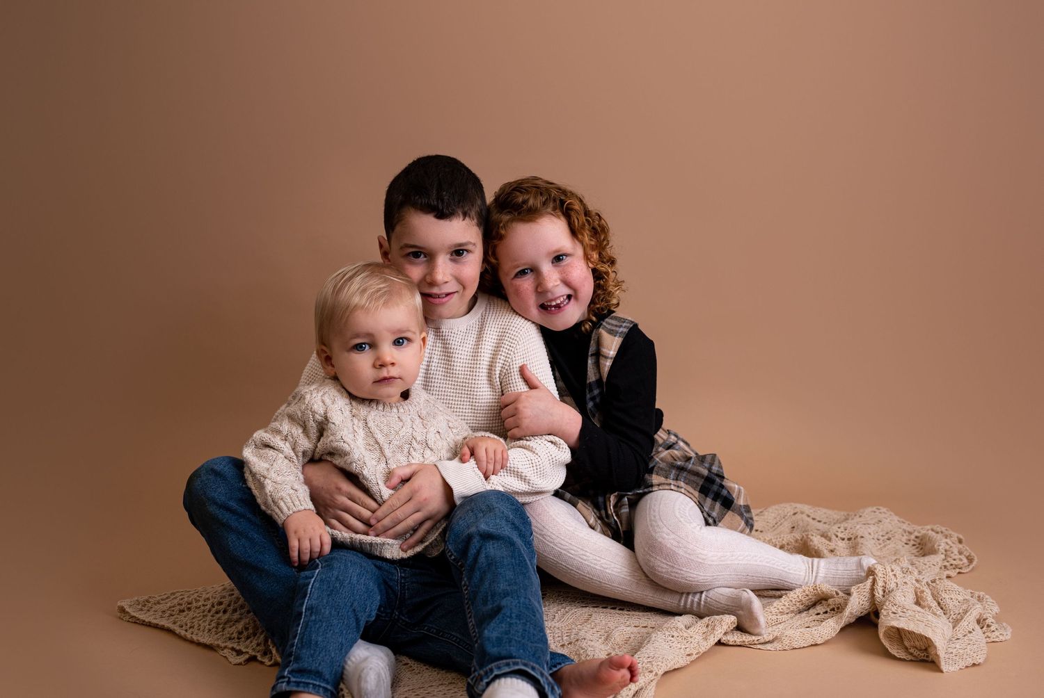Three siblings sit together against a brown backdrop wearing casual clothing and sharing a sweet moment.