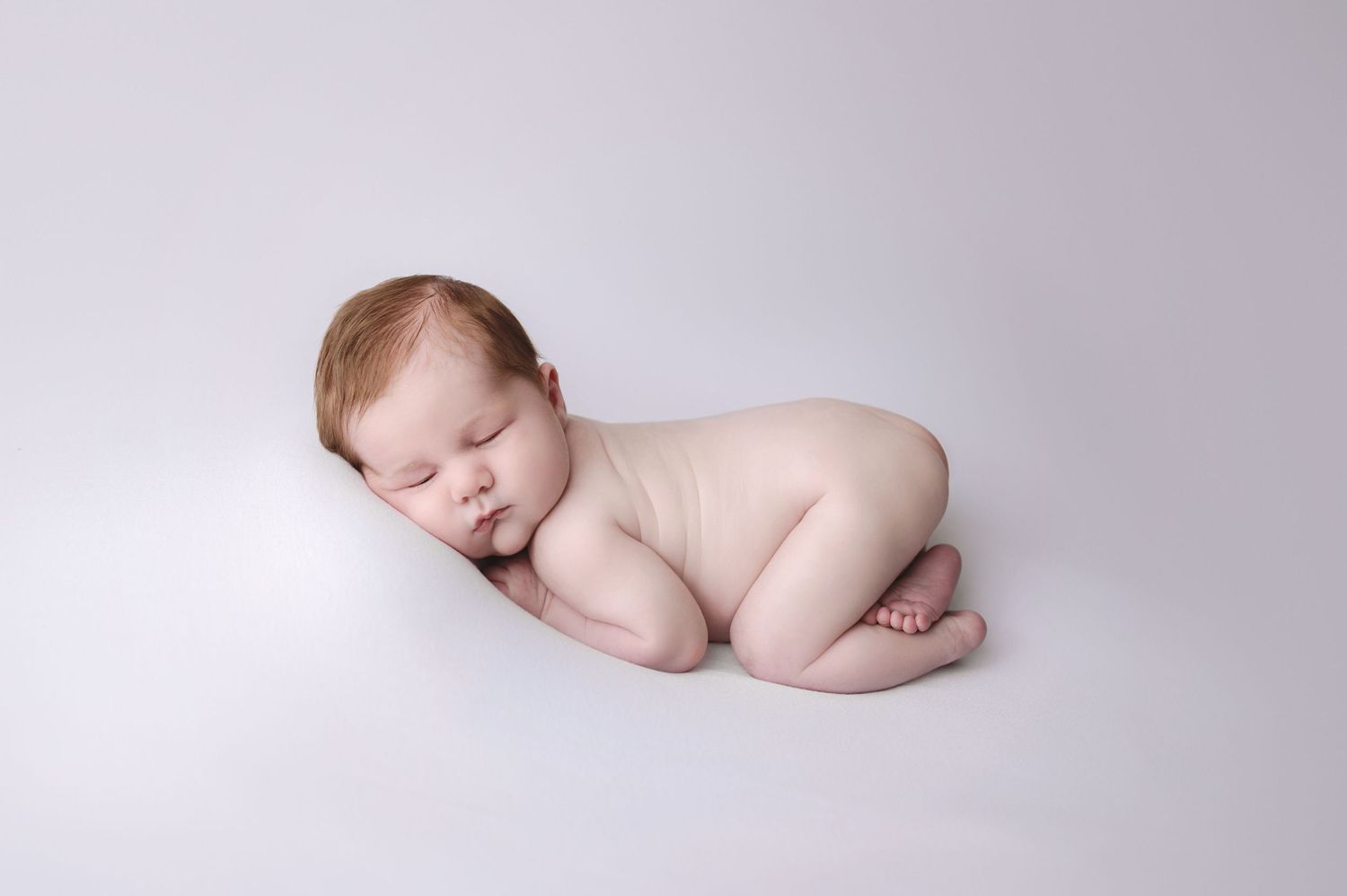 Peaceful sleeping newborn baby curled up in soft white studio lighting against neutral background.