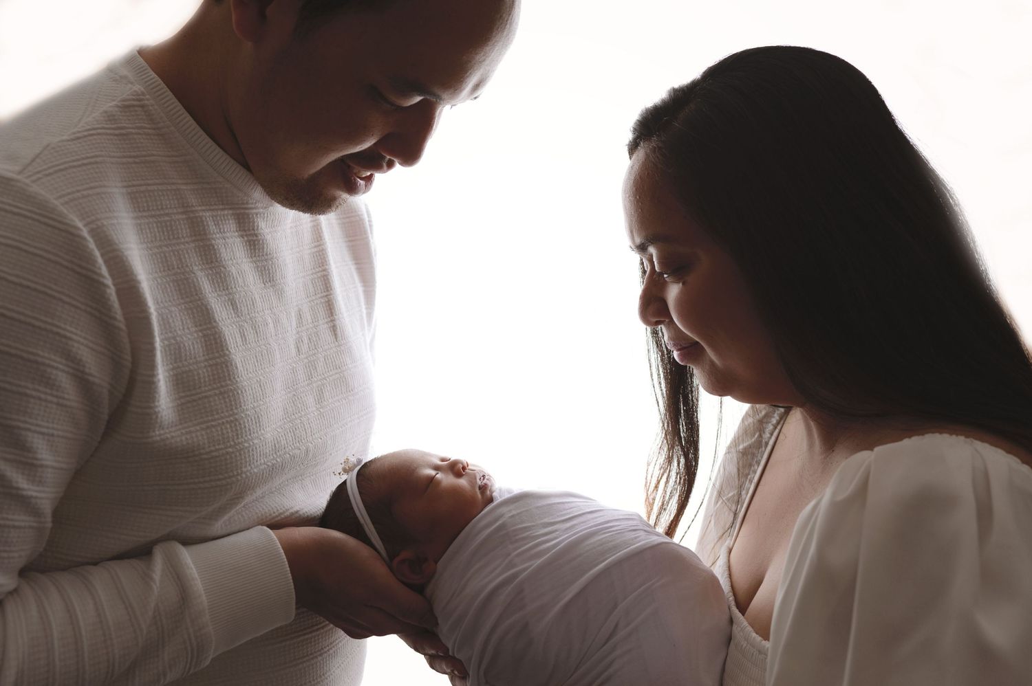Parents gently cradling their newborn baby in a tender moment silhouetted against soft window light.
