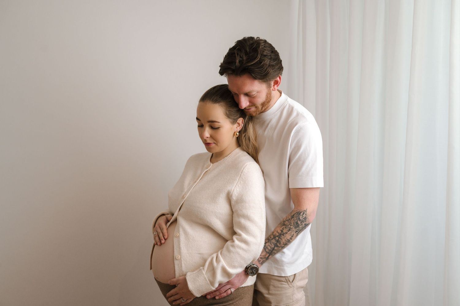 Couple tenderly cradling pregnant belly during maternity photo shoot against white background.