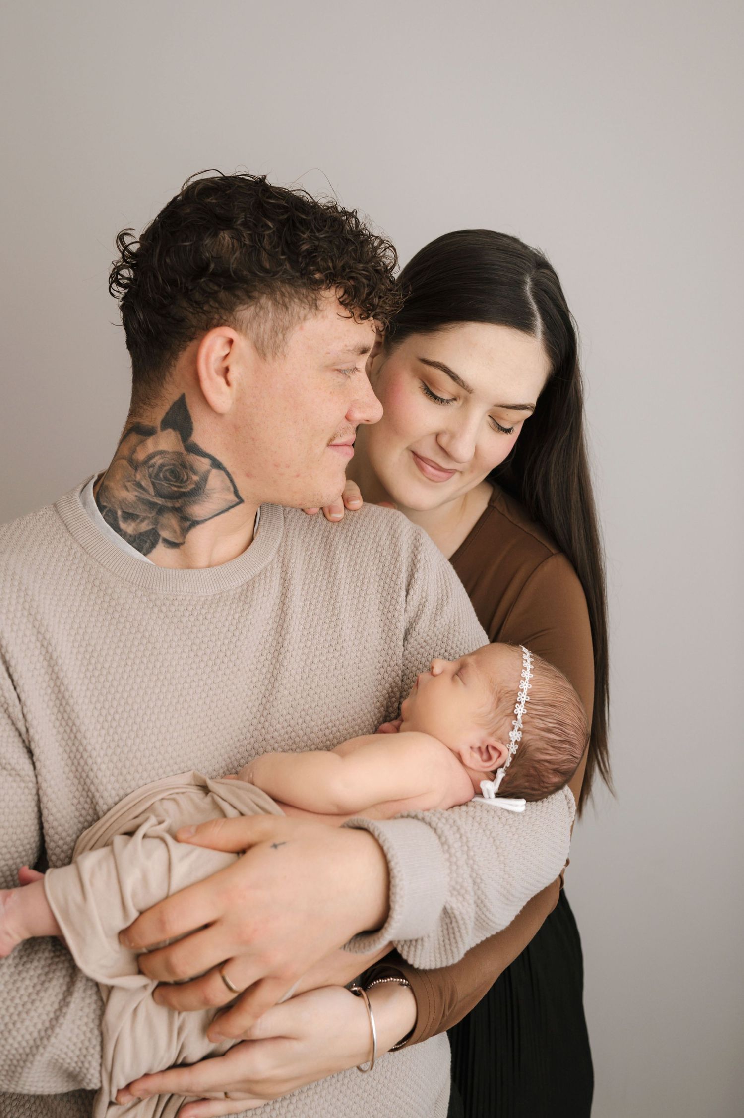 A young couple with tattoos tenderly holds their sleeping newborn baby in a soft, neutral-toned studio portrait.