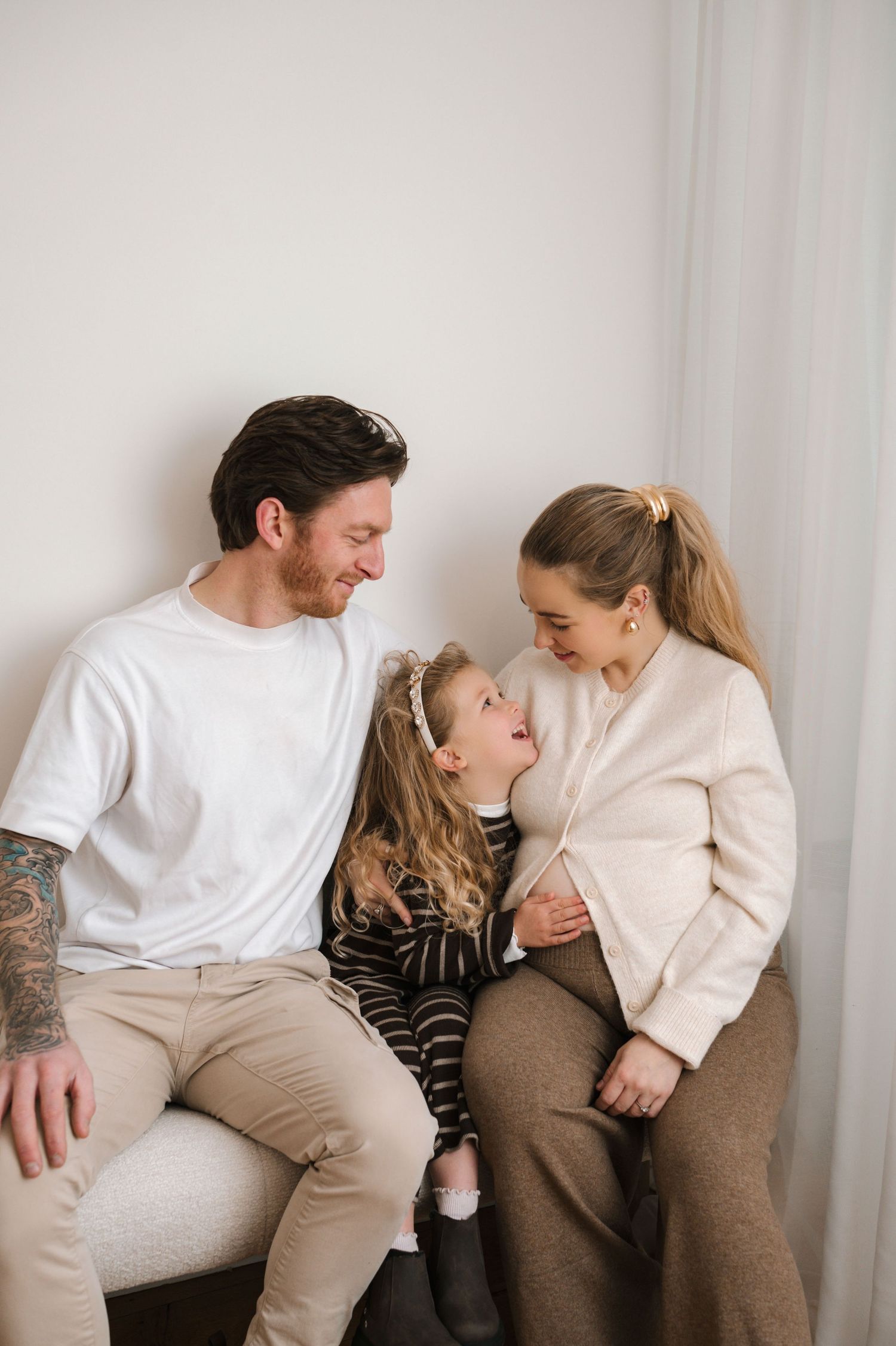 A happy family of three sits together, with a young girl between her smiling parents in a bright, cozy room.