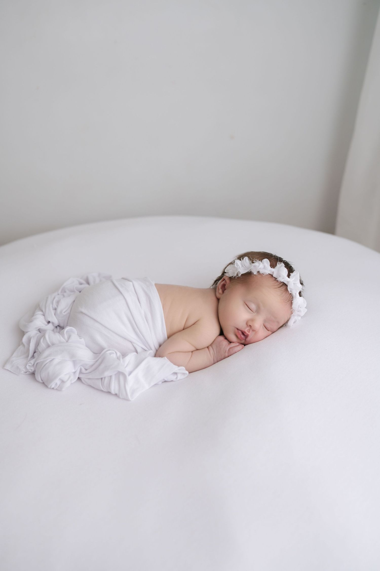 Sleeping newborn baby in white swaddle lying on soft white bedding in minimalist studio setting.