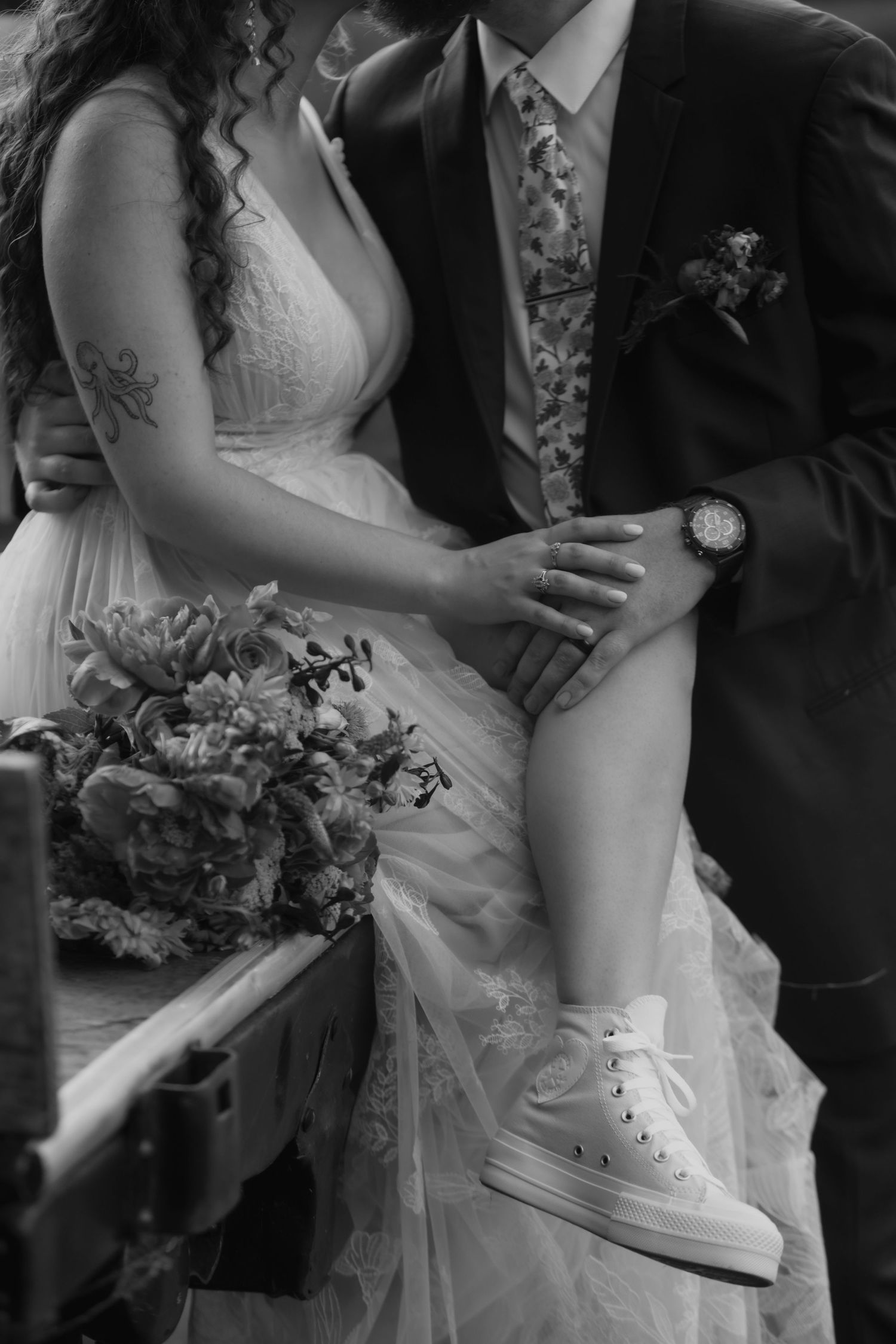 Black and white wedding photo showing bride in white sneakers and wedding dress with groom in suit holding bouquet.