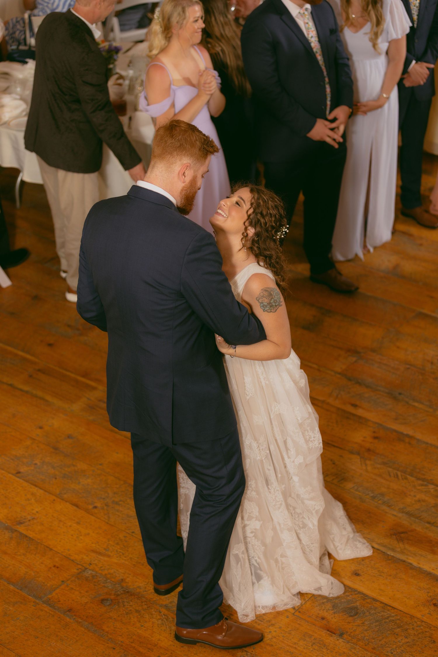 A couple shares an intimate dance at their wedding reception on a rustic wooden floor while guests look on.