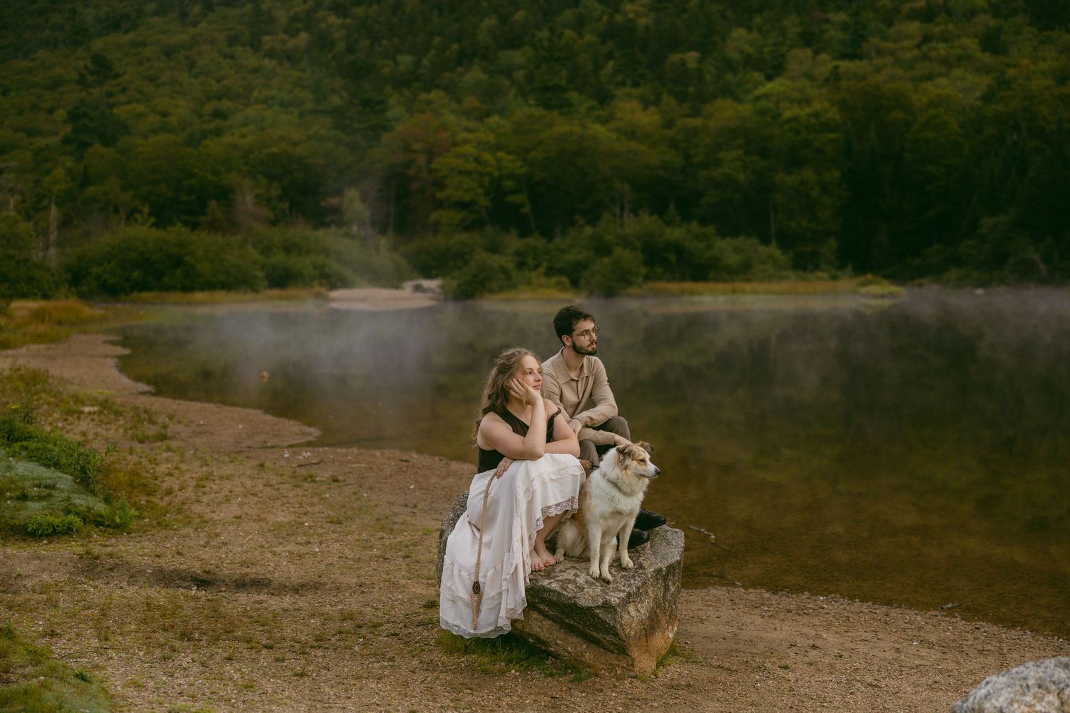 A couple walks with their golden retriever dog along a scenic wooded path at sunset during an engagement photoshoot.