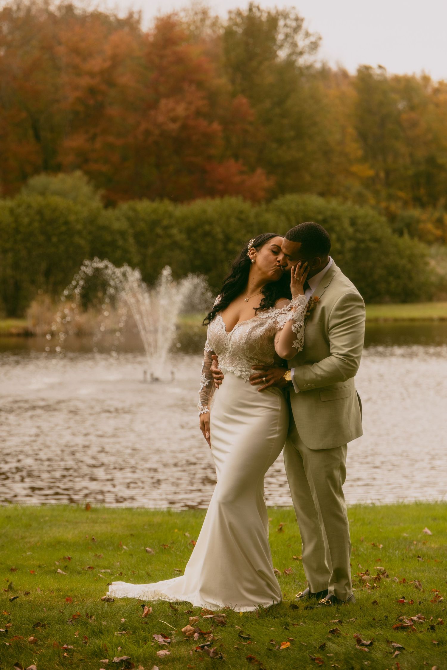 A couple in wedding attire share a romantic moment by a fountain in a park with autumn foliage in the background.