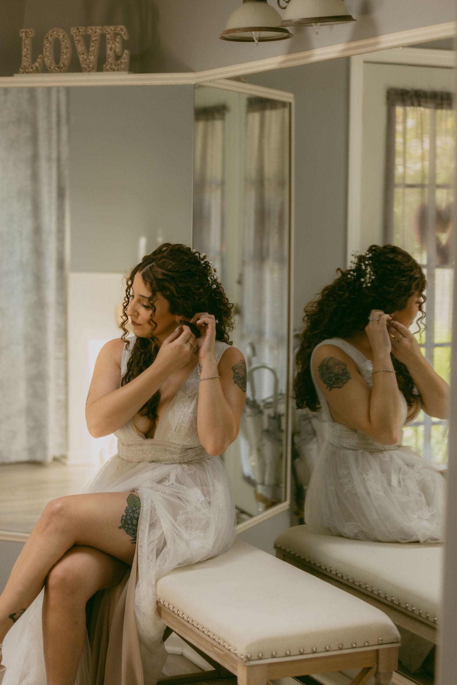A woman in a white dress adjusts her jewelry while sitting at a vanity mirror in a softly lit room.