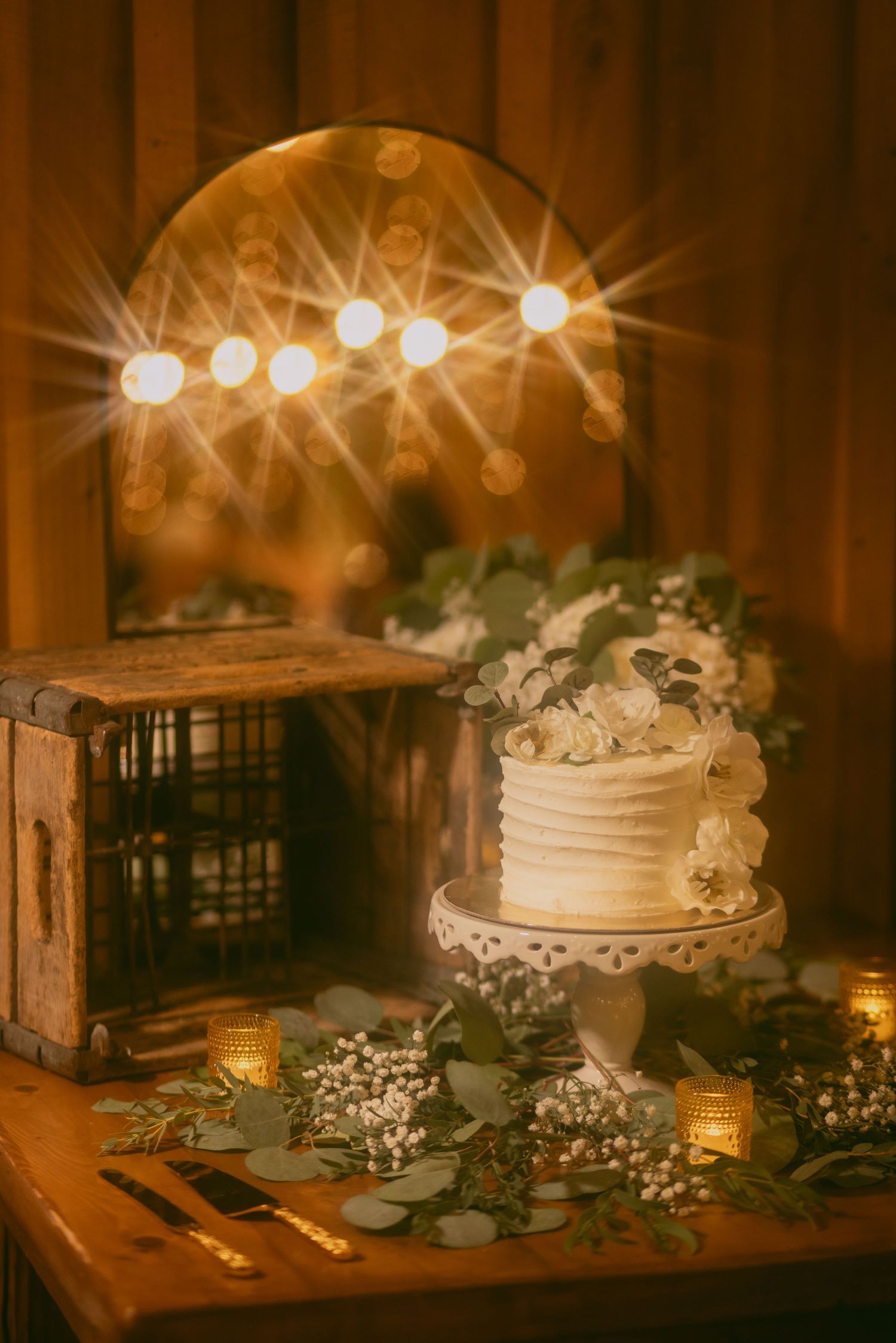 A rustic wedding cake display with glowing candles and baby's breath flowers on a wooden table in warm ambient lighting.