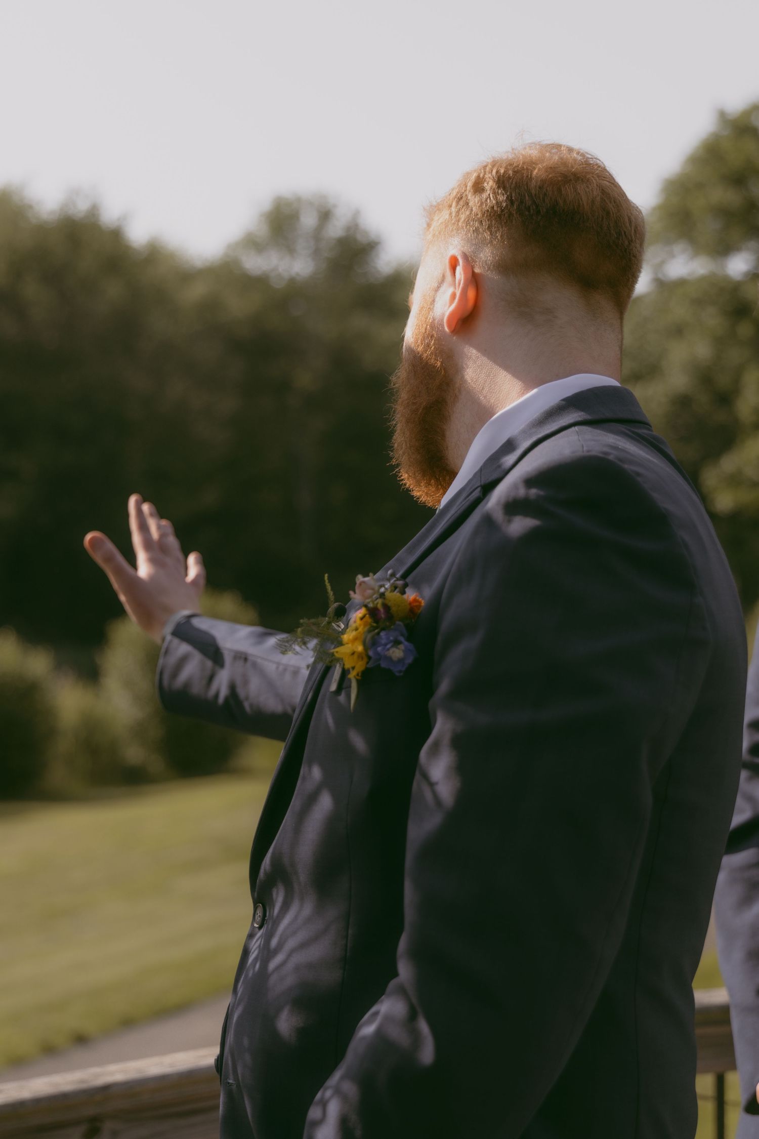 A groom in a navy suit with sunflower boutonniere gestures with his hand while standing outdoors on a sunny day.