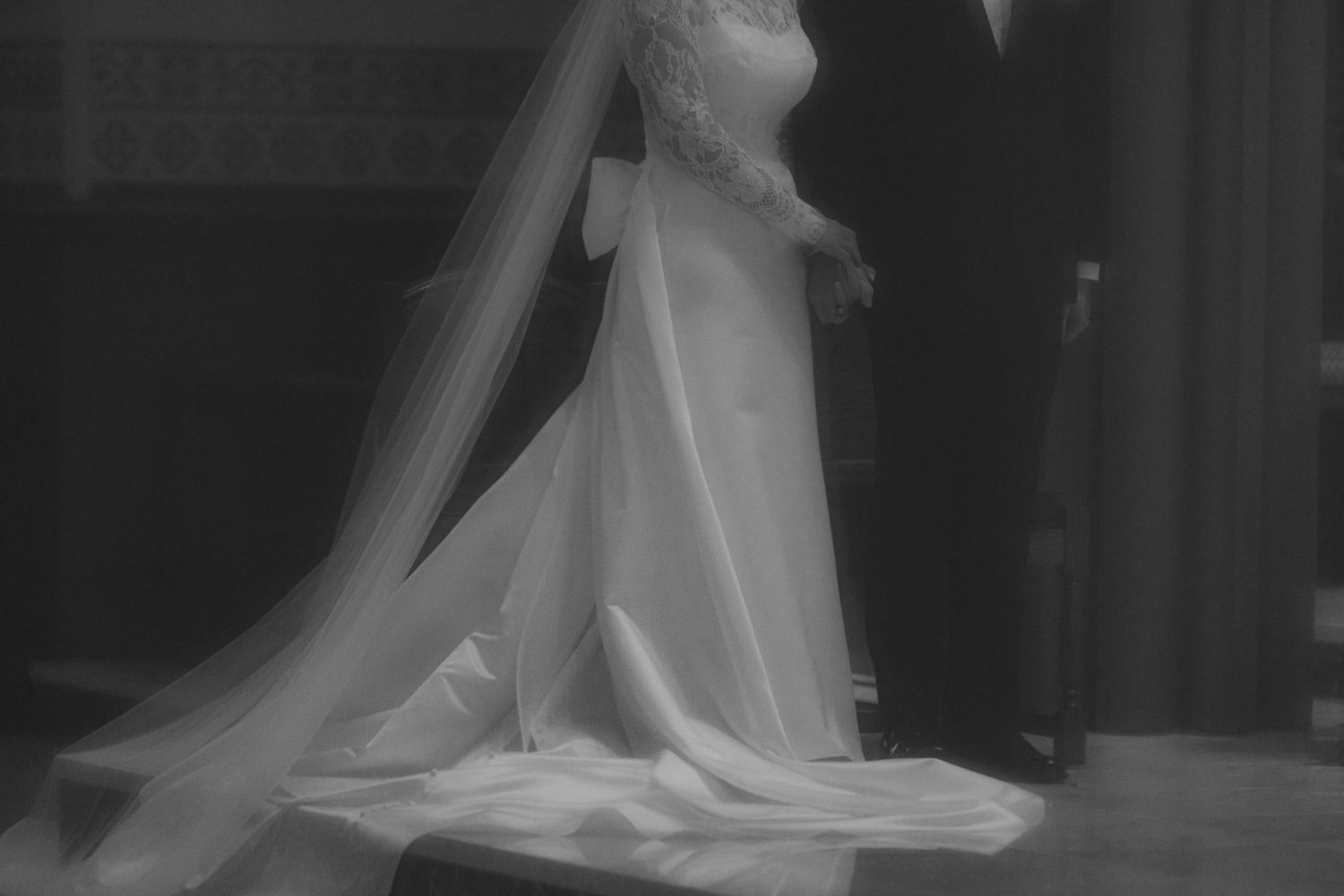 A black and white photograph of a wedding dress with a long flowing train and veil in soft focus.