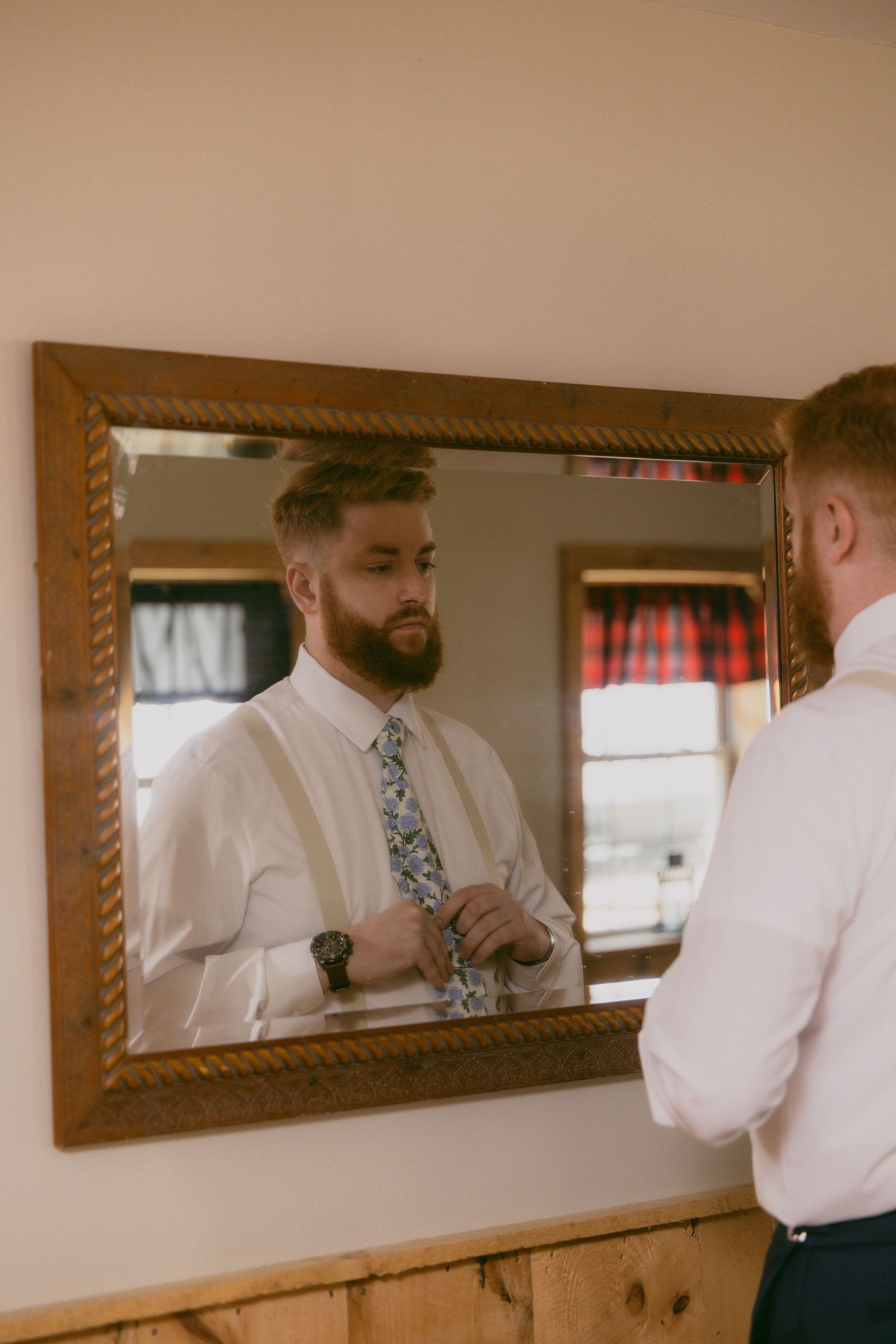 A person wearing a white dress shirt and floral tie adjusts their outfit while looking in a wooden framed mirror.