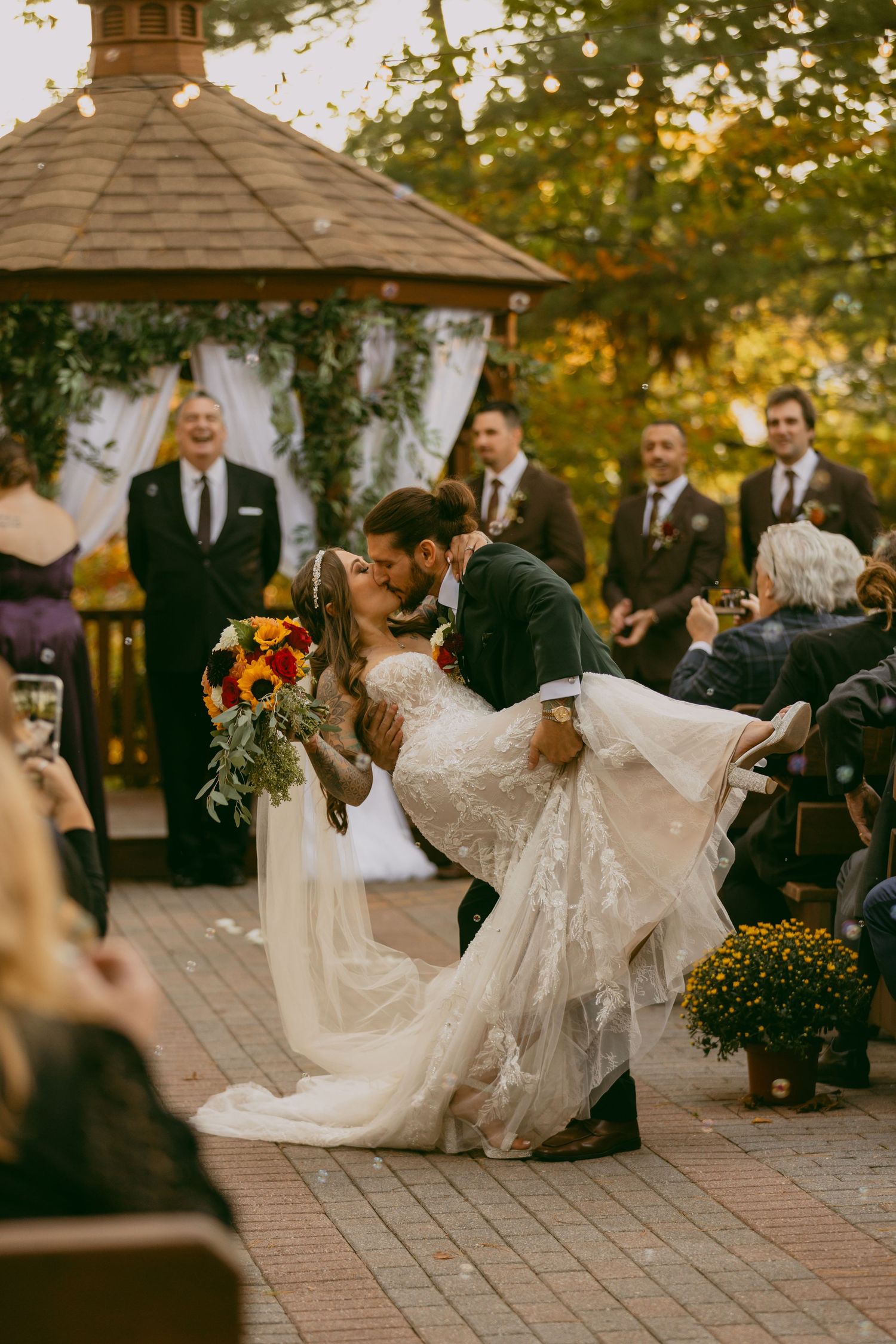 A romantic wedding couple dances together on a stone patio during an autumn outdoor reception at sunset.