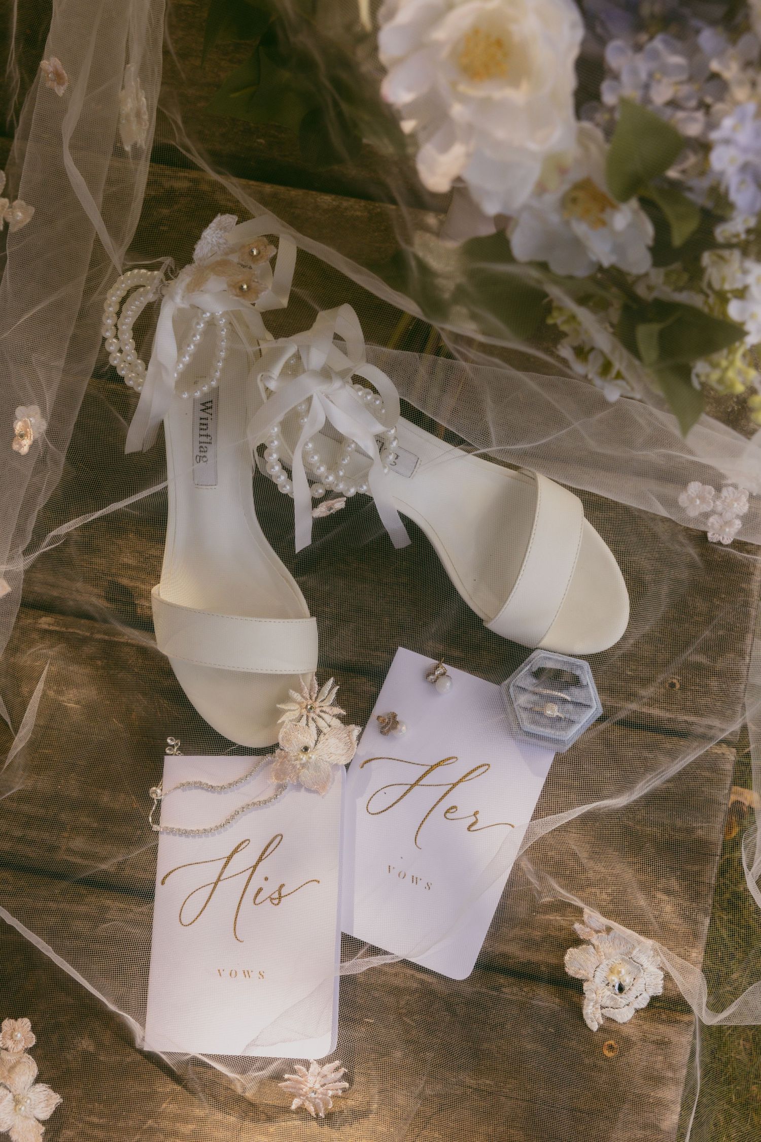 Elegant white bridal heels and 'His' wedding vow cards arranged with delicate flowers and veil on wooden surface.