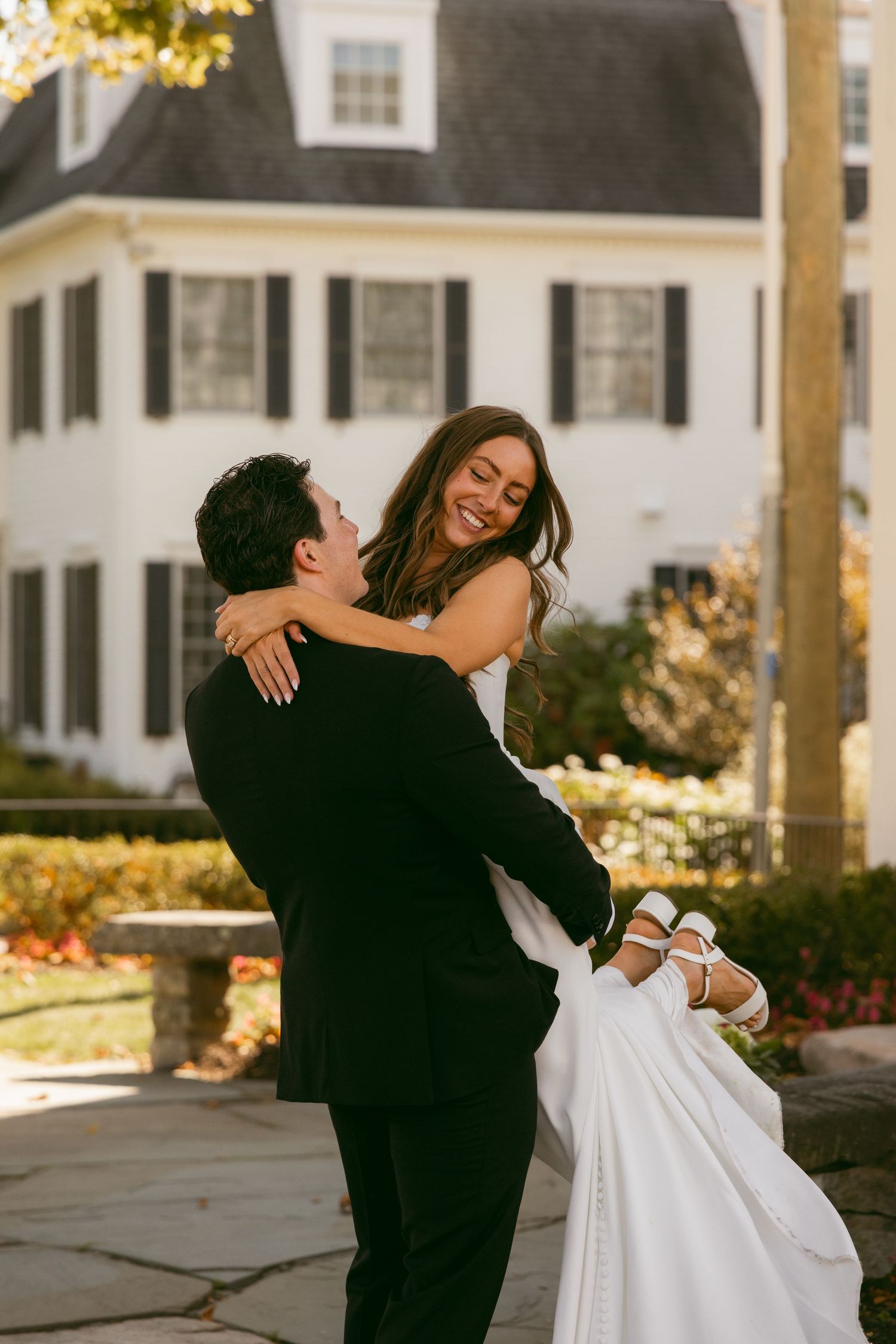 A couple in formal wedding attire dances and embraces outdoors near a white house during golden hour.