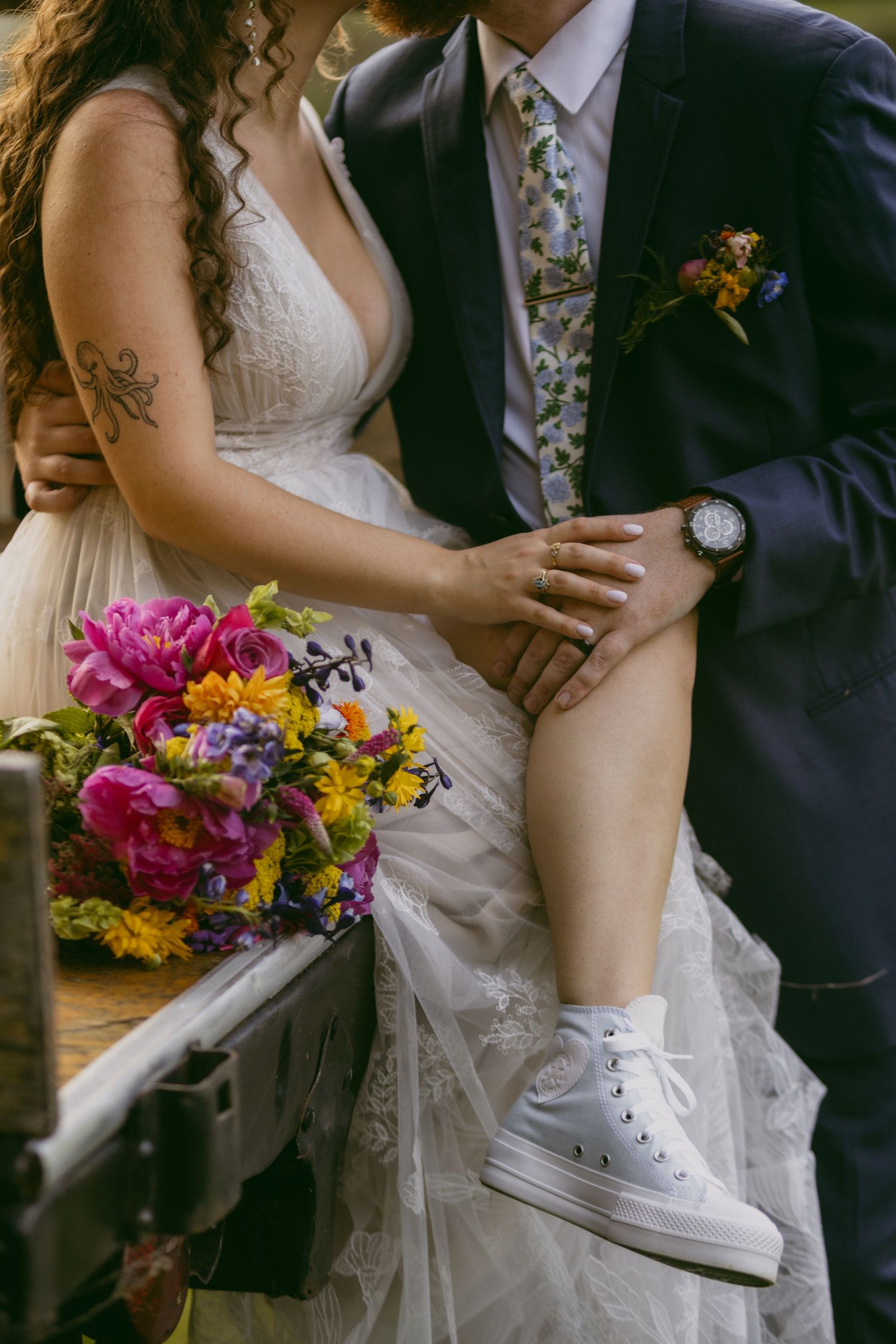 Close-up of bride in white dress and groom in suit with colorful bouquet and white sneakers visible.