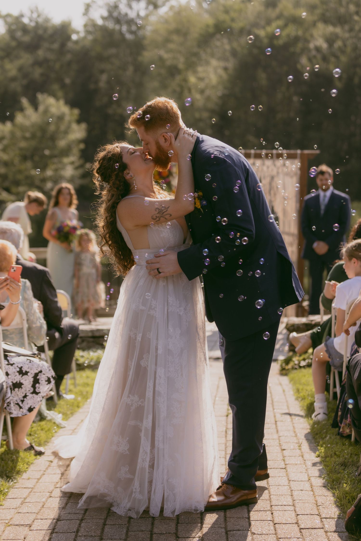 A romantic outdoor wedding ceremony with soap bubbles floating in the sunlit air as a couple shares their first kiss on a brick path.