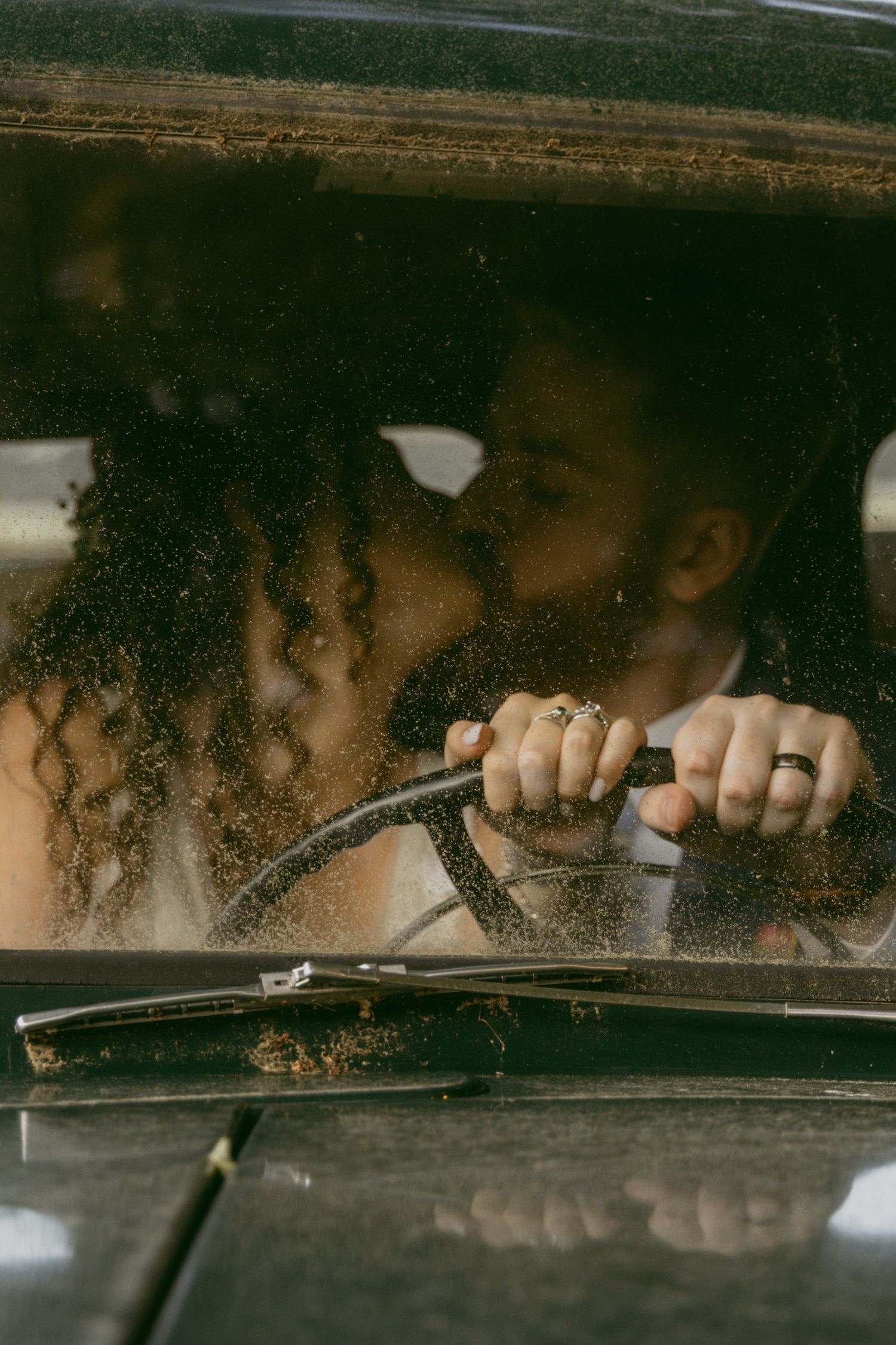 Two people share an intimate moment through a car windshield with raindrops and soft natural lighting.