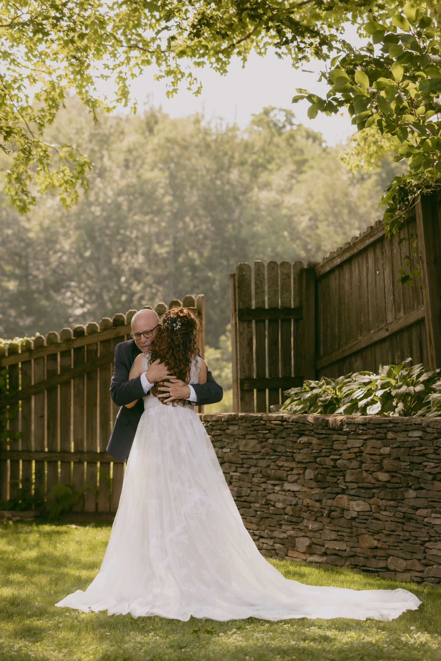 A romantic garden wedding scene with a flowing white gown against a rustic wooden fence and stone wall backdrop.
