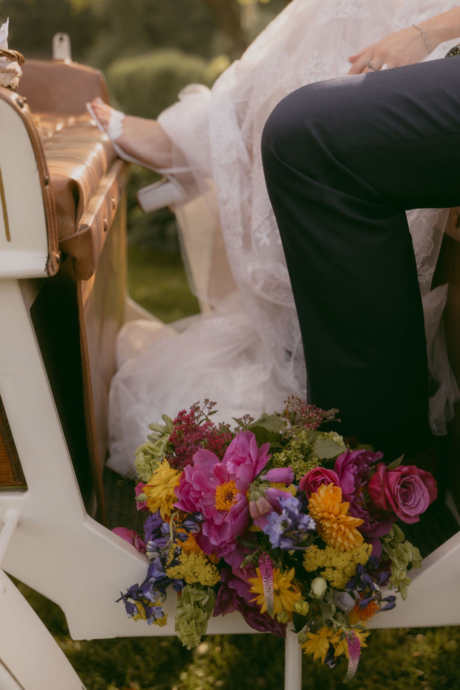 Two people in wedding attire sit together in front of colorful spring flowers and wooden decor letters at an outdoor ceremony.