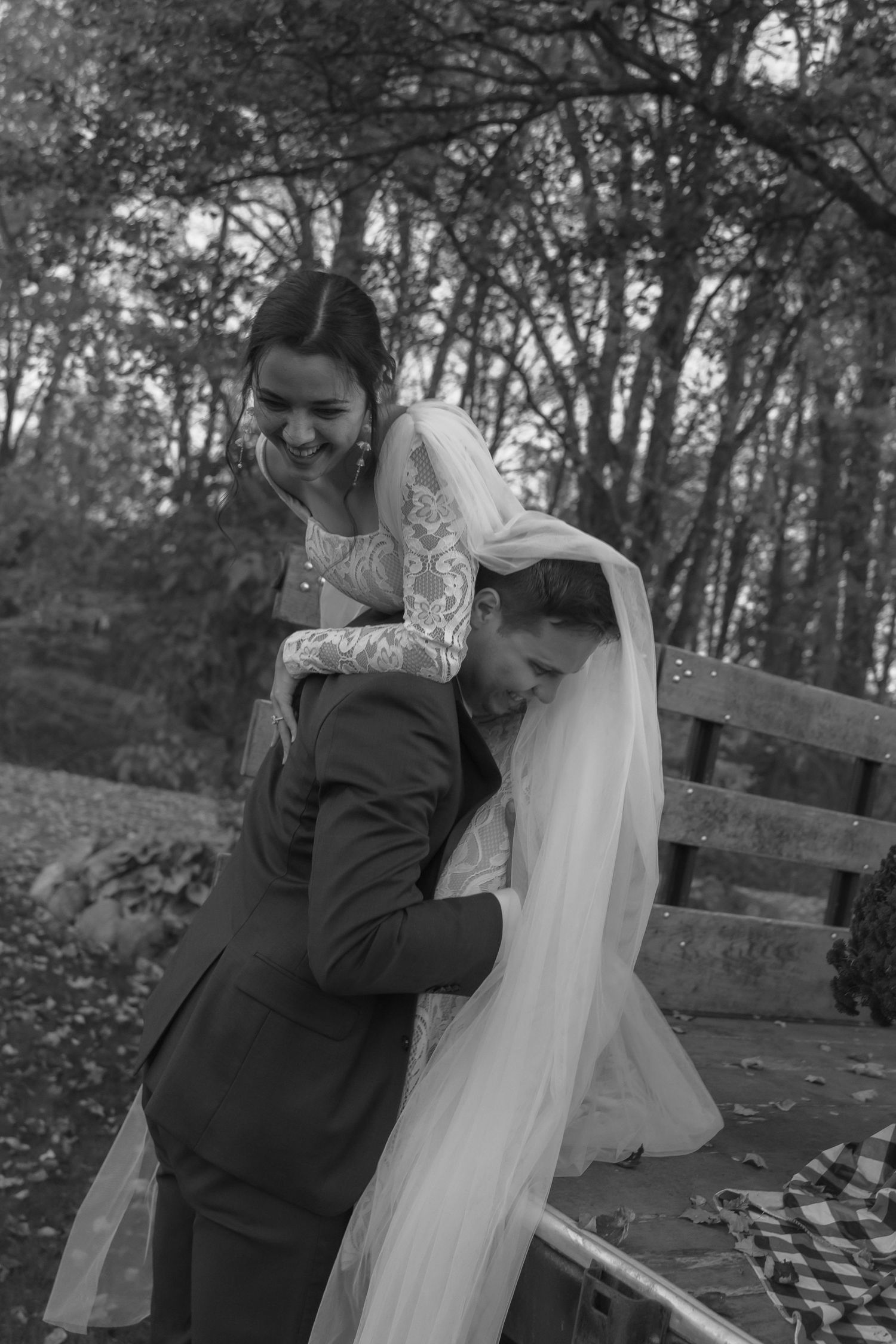 A black and white wedding portrait shows a romantic moment between newlyweds on a wooden bench surrounded by trees.