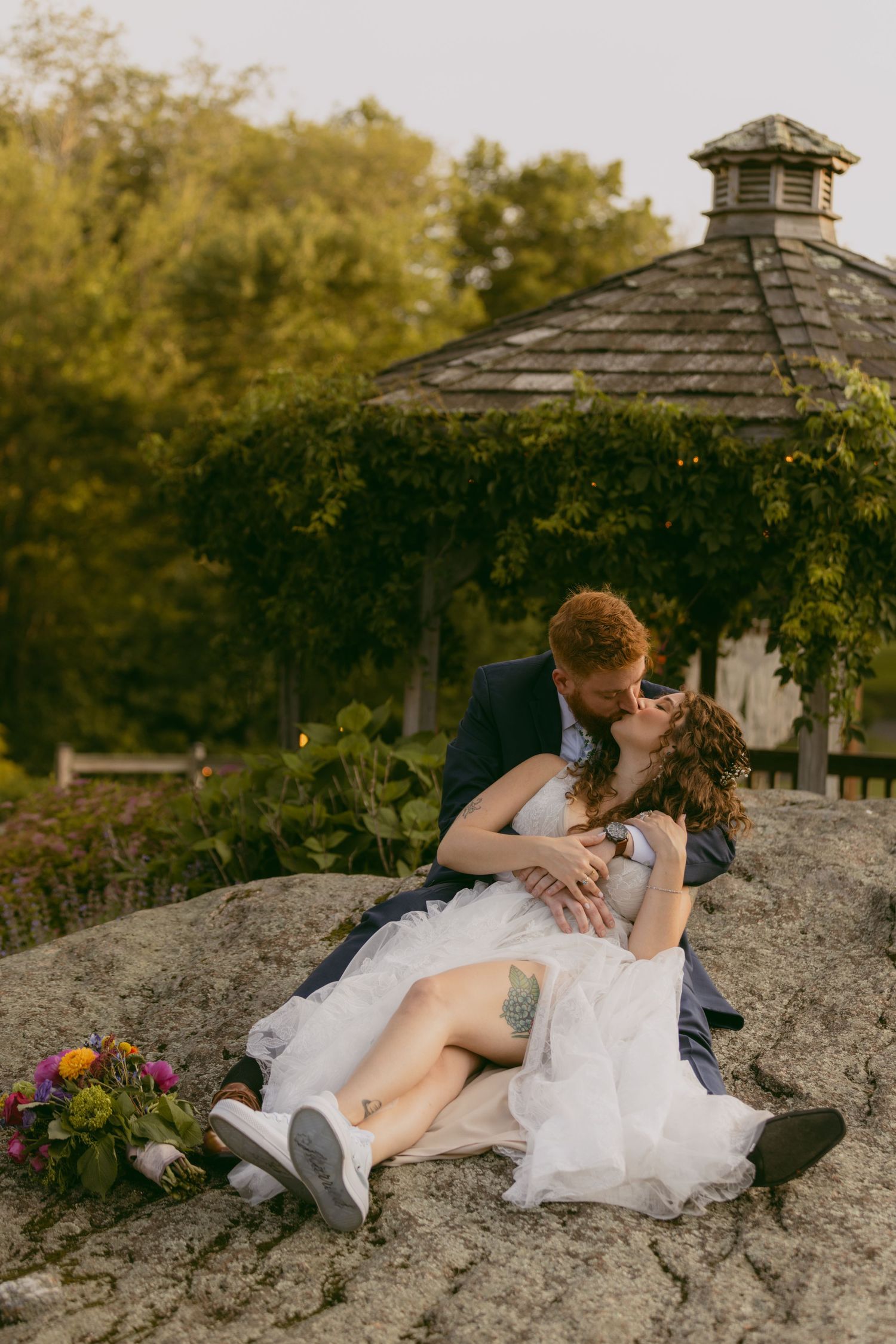 A romantic couple in wedding attire share an intimate moment on a rocky outcrop near a garden gazebo.